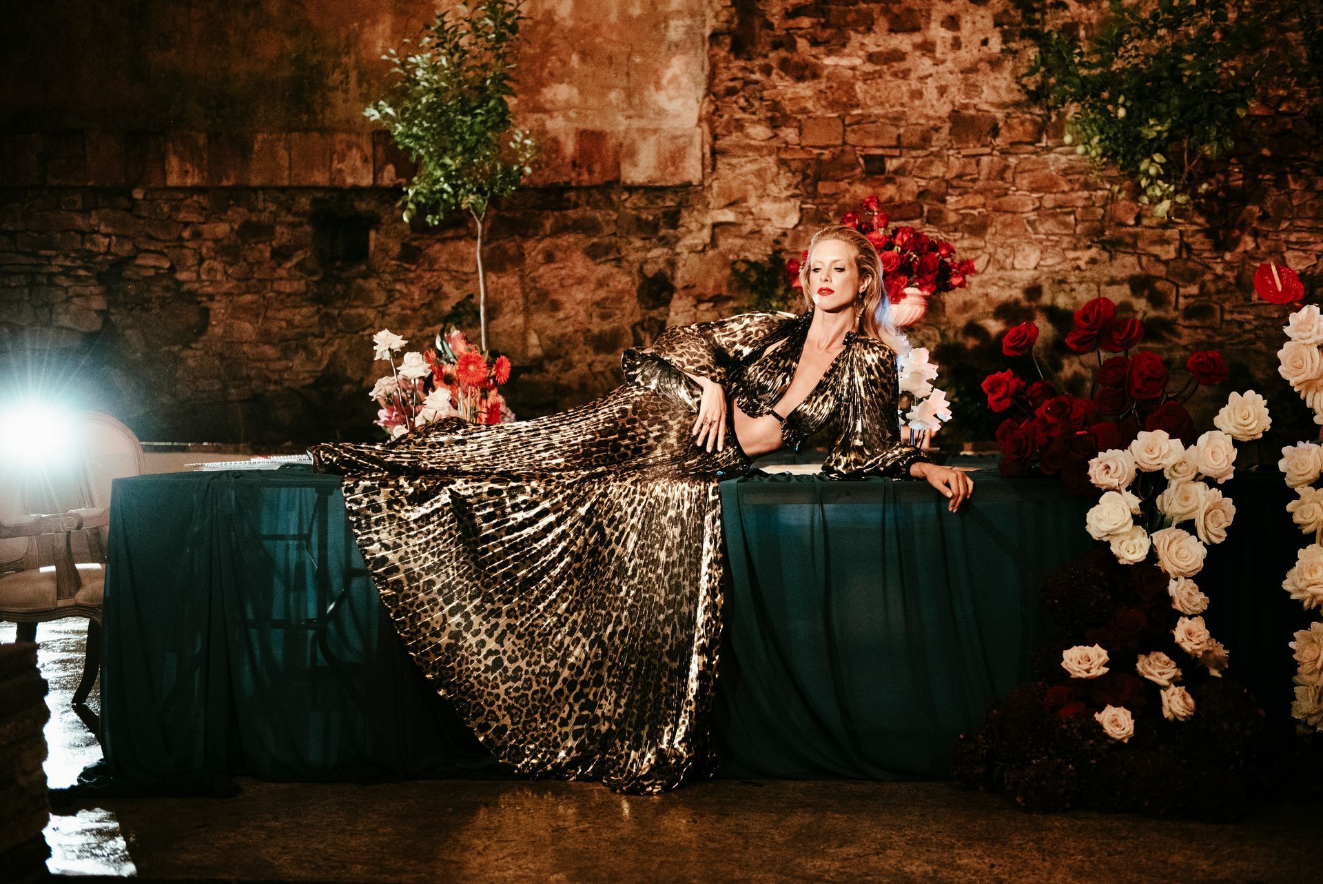 Woman in gold gown reclines on a teal-draped table amid red and white flowers against a rustic brick wall.