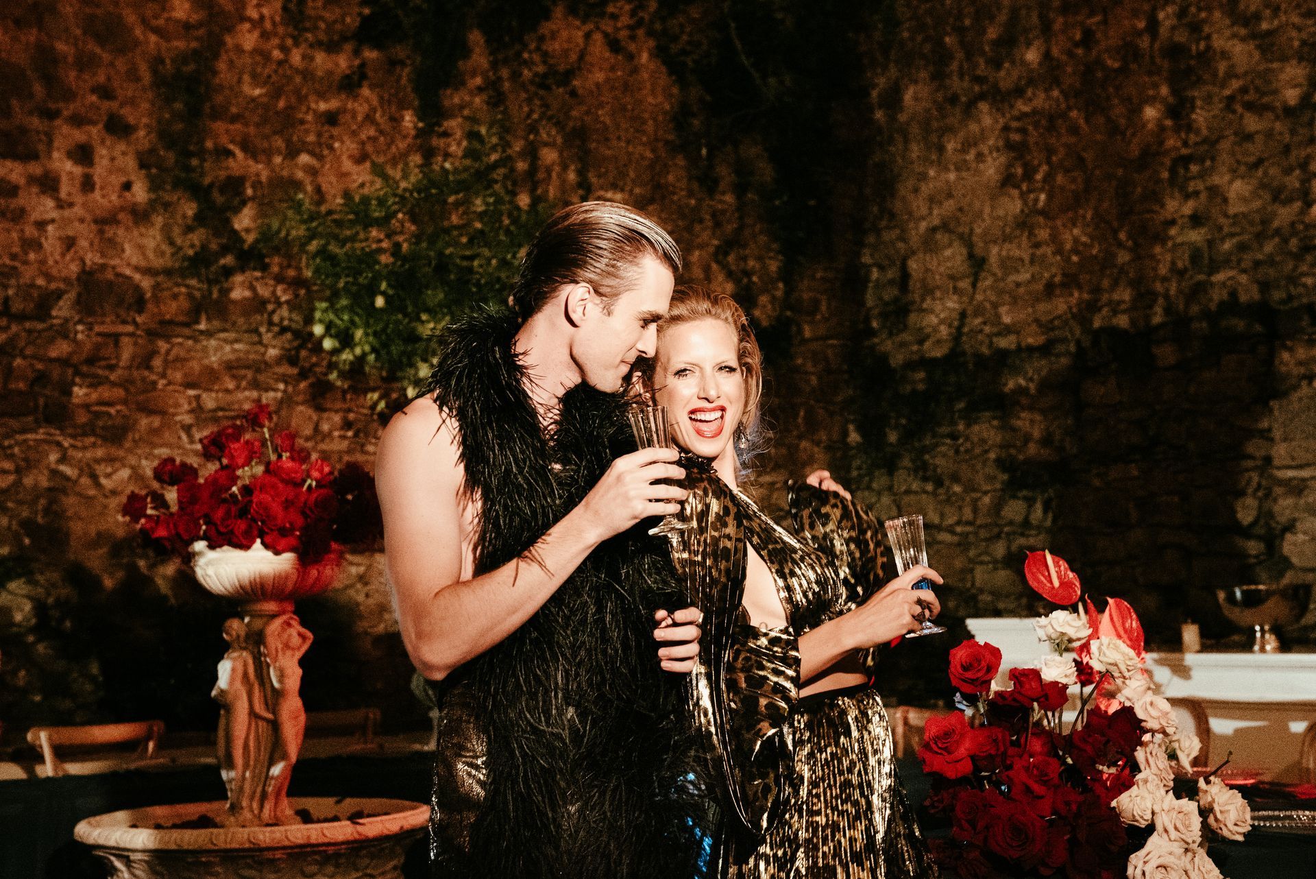 Couple in metallic outfits, toasting champagne, outdoors near red flowers and a fountain.