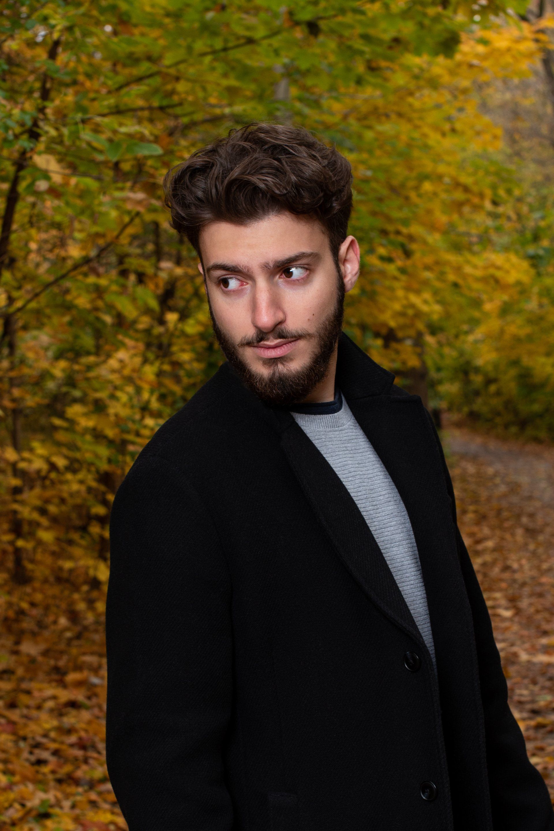 Man in black coat, looking over shoulder, standing in autumn forest with yellow leaves.