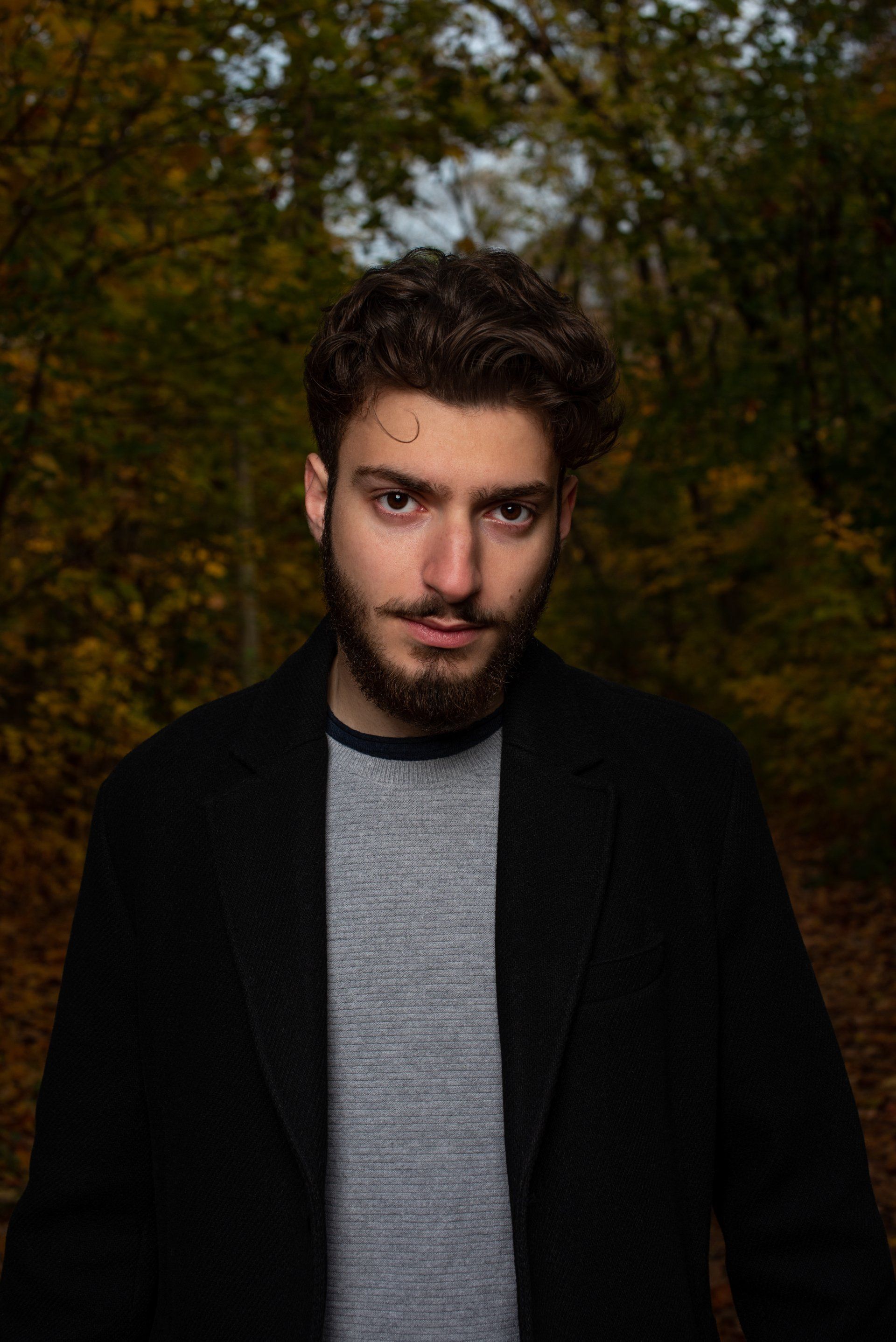 Man with beard in a black coat and gray sweater, standing in a forest with autumn foliage.