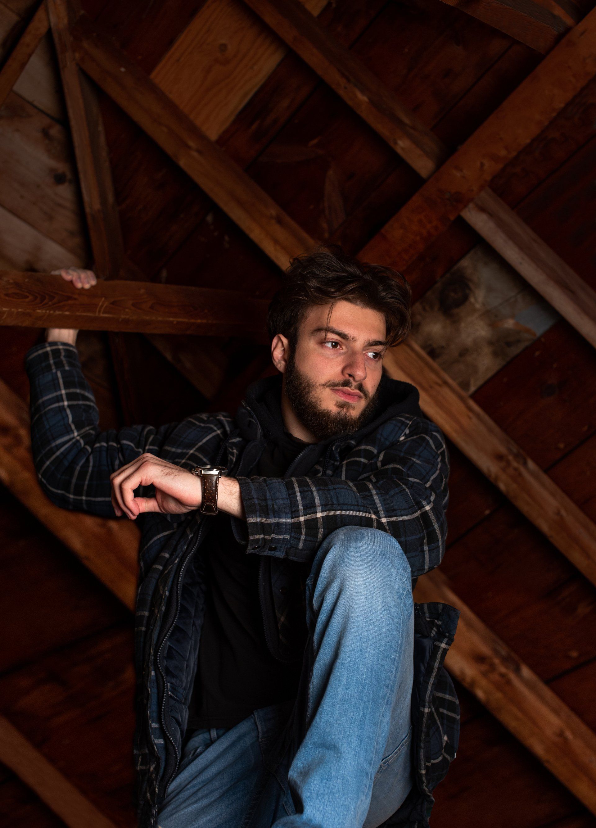 Man in jeans and flannel shirt in an attic, looking to the side, leaning on wooden beams.