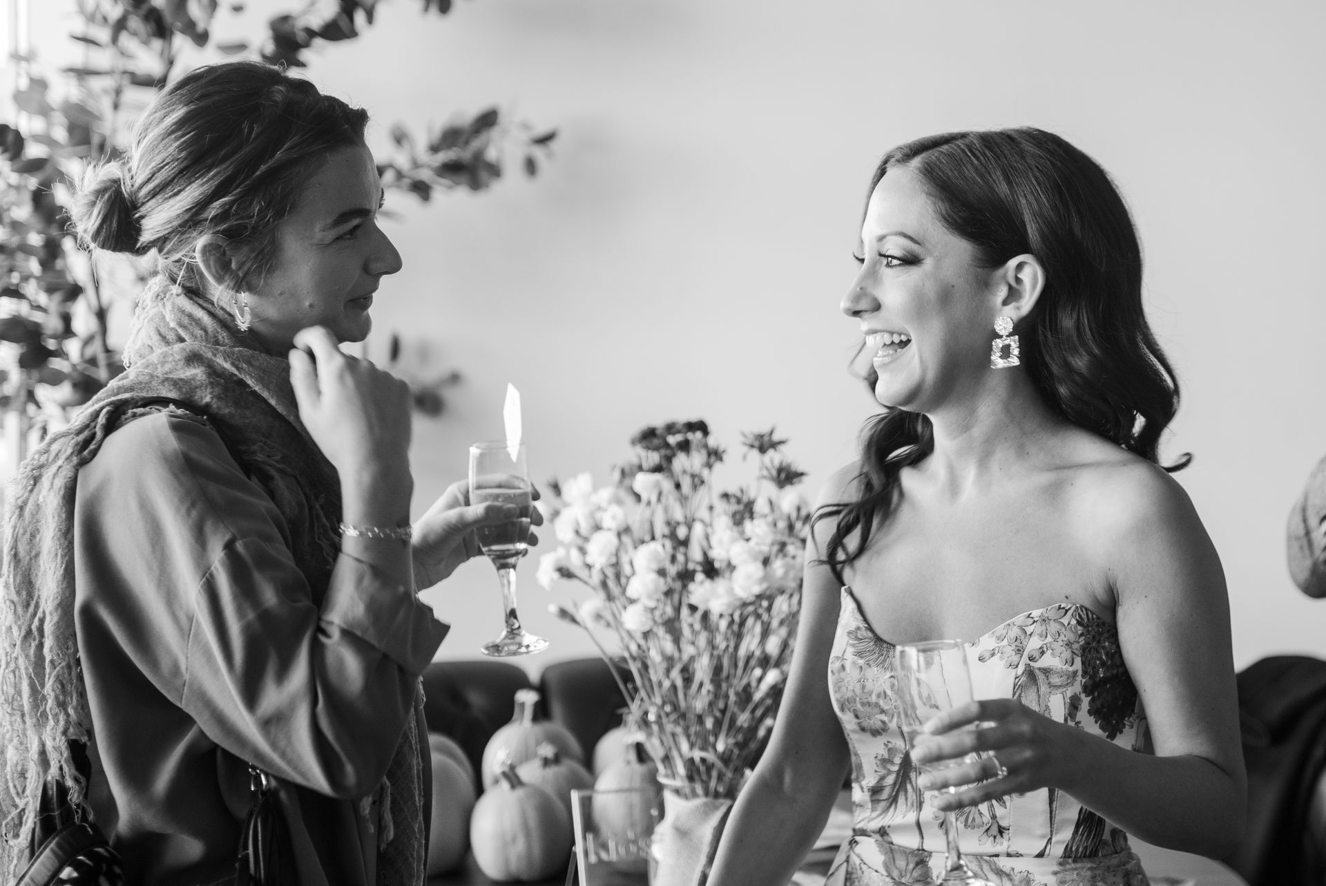 Two women laughing, one holding a drink. Florals and pumpkins in background.