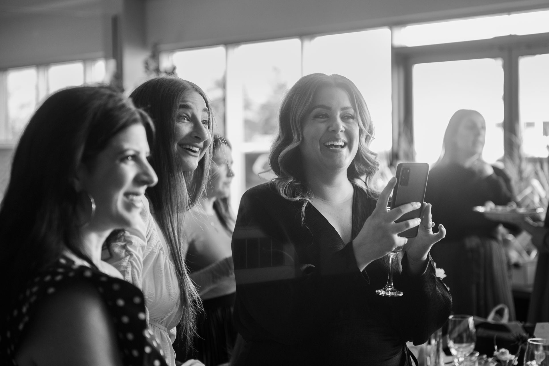 Women smile and laugh while one takes a photo at an indoor event with a bright window backdrop.