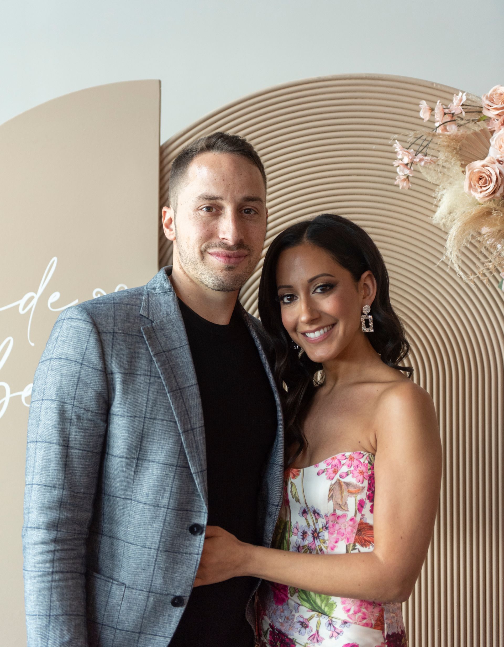 Couple smiling, posing in front of a tan archway decorated with flowers. Man in blazer, woman in floral dress.