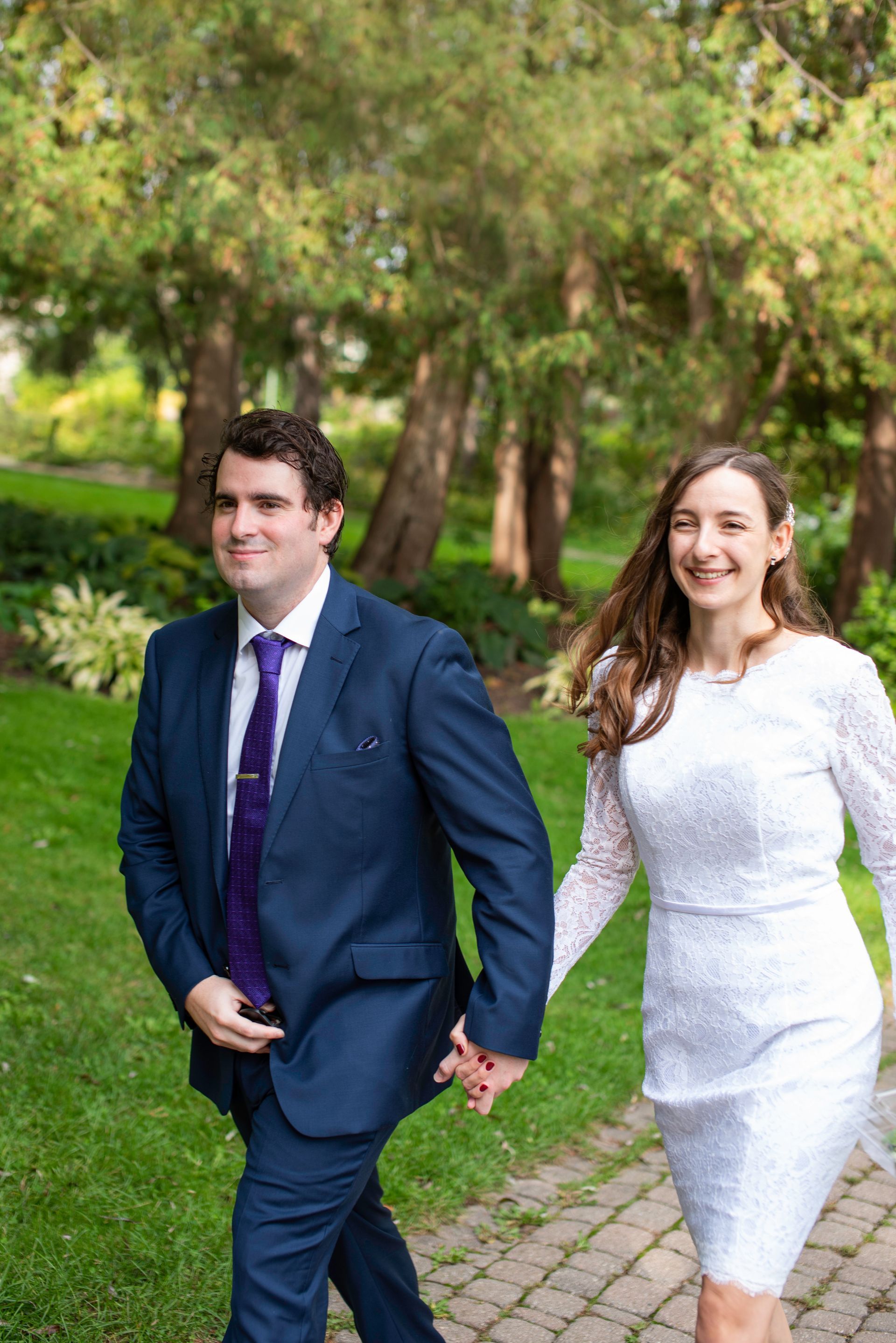 Couple holding hands, walking on a path in a park; the man in a blue suit, the woman in a white dress.