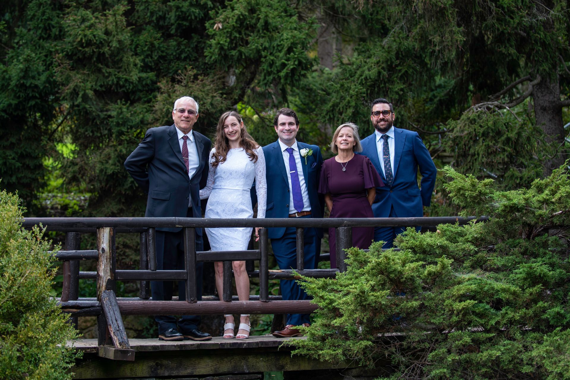 Group of people on a wooden bridge, possibly a wedding photo. Green foliage in the background.