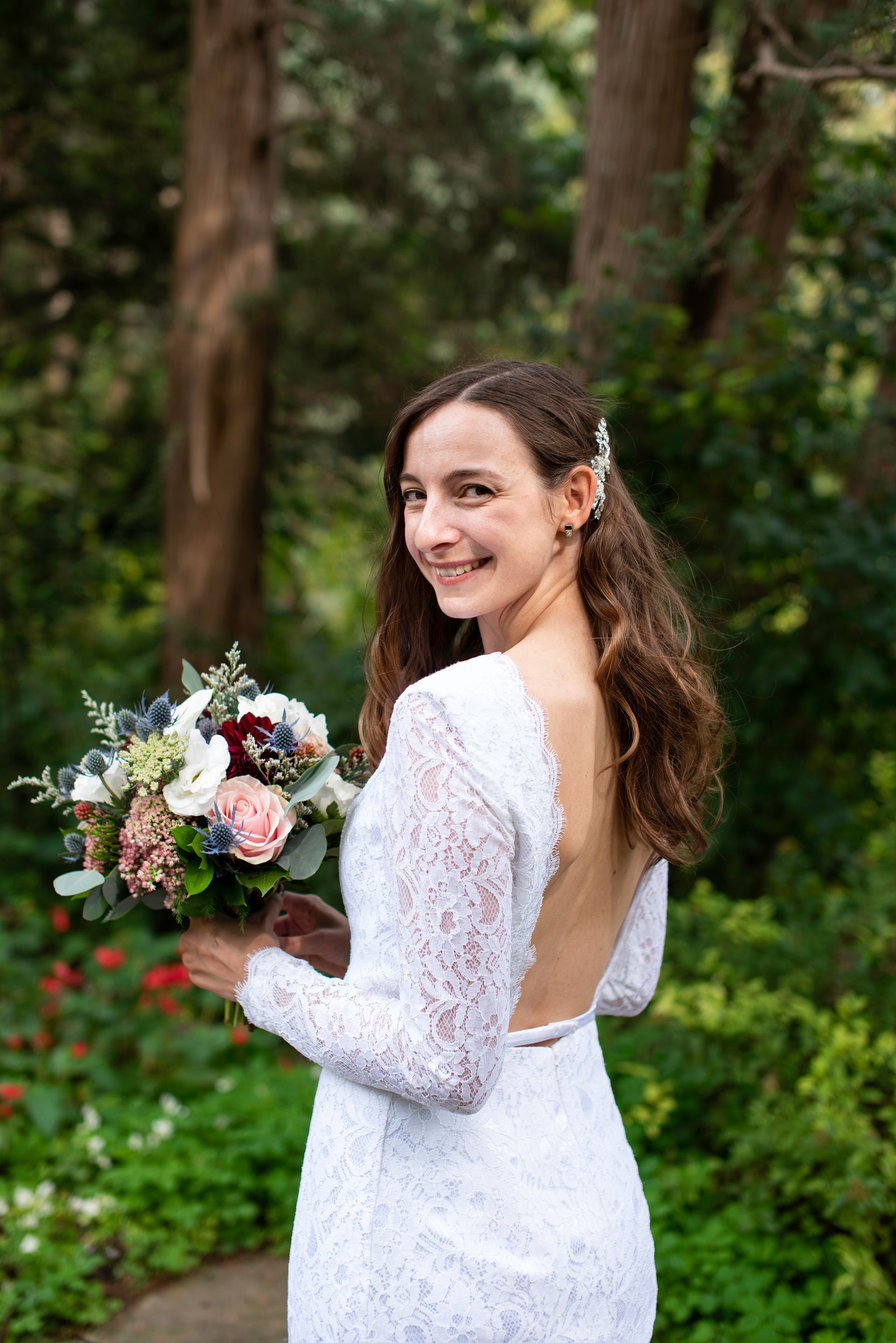 Bride in white lace dress, holding bouquet, looking over shoulder, smiling in a garden.