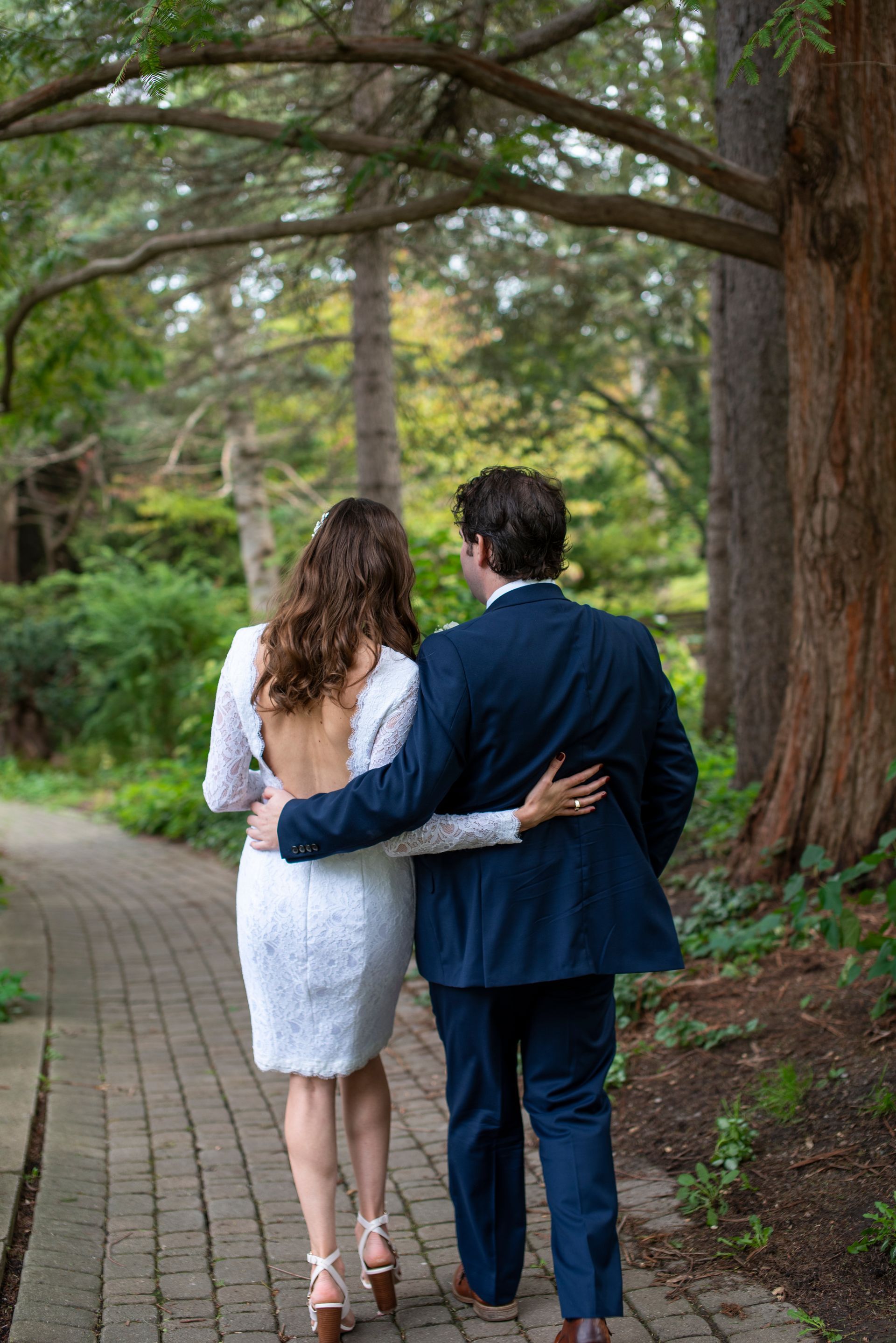 Couple walking on a brick path through a wooded area; woman in white dress, man in blue suit.