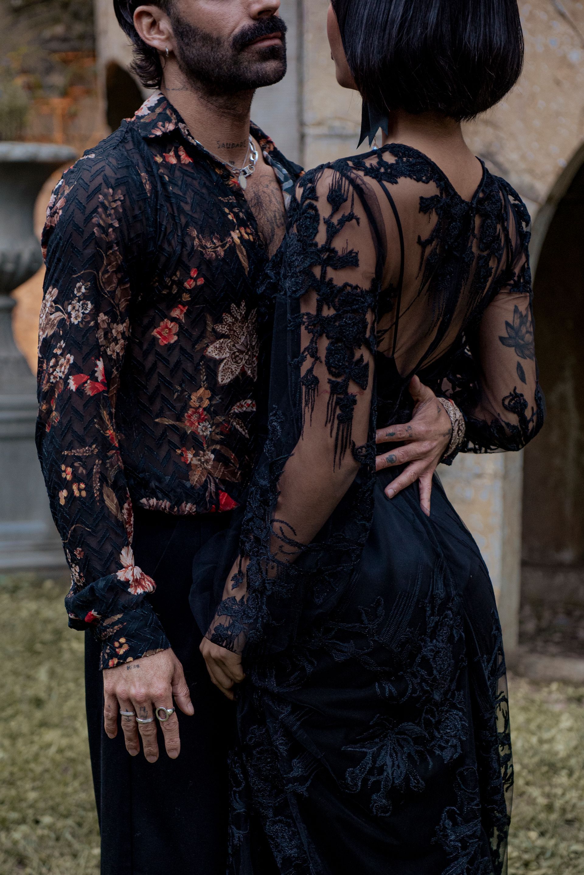Man in floral shirt embraces person in black lace dress outdoors.