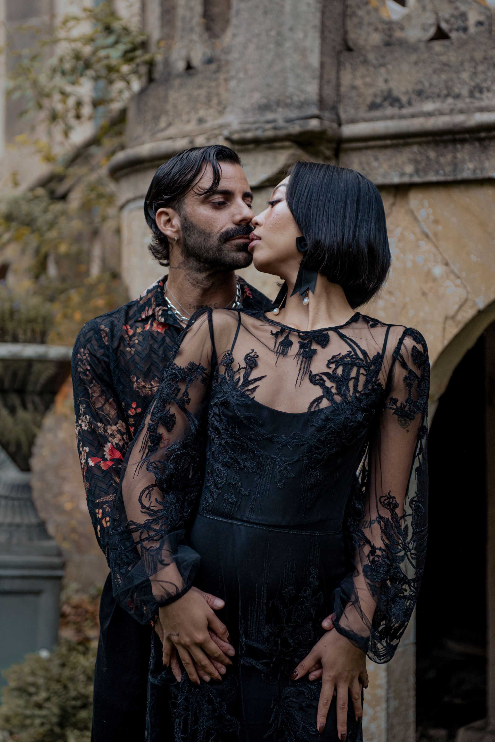 Couple embracing, about to kiss. Man in floral shirt, woman in black lace dress, standing outside.