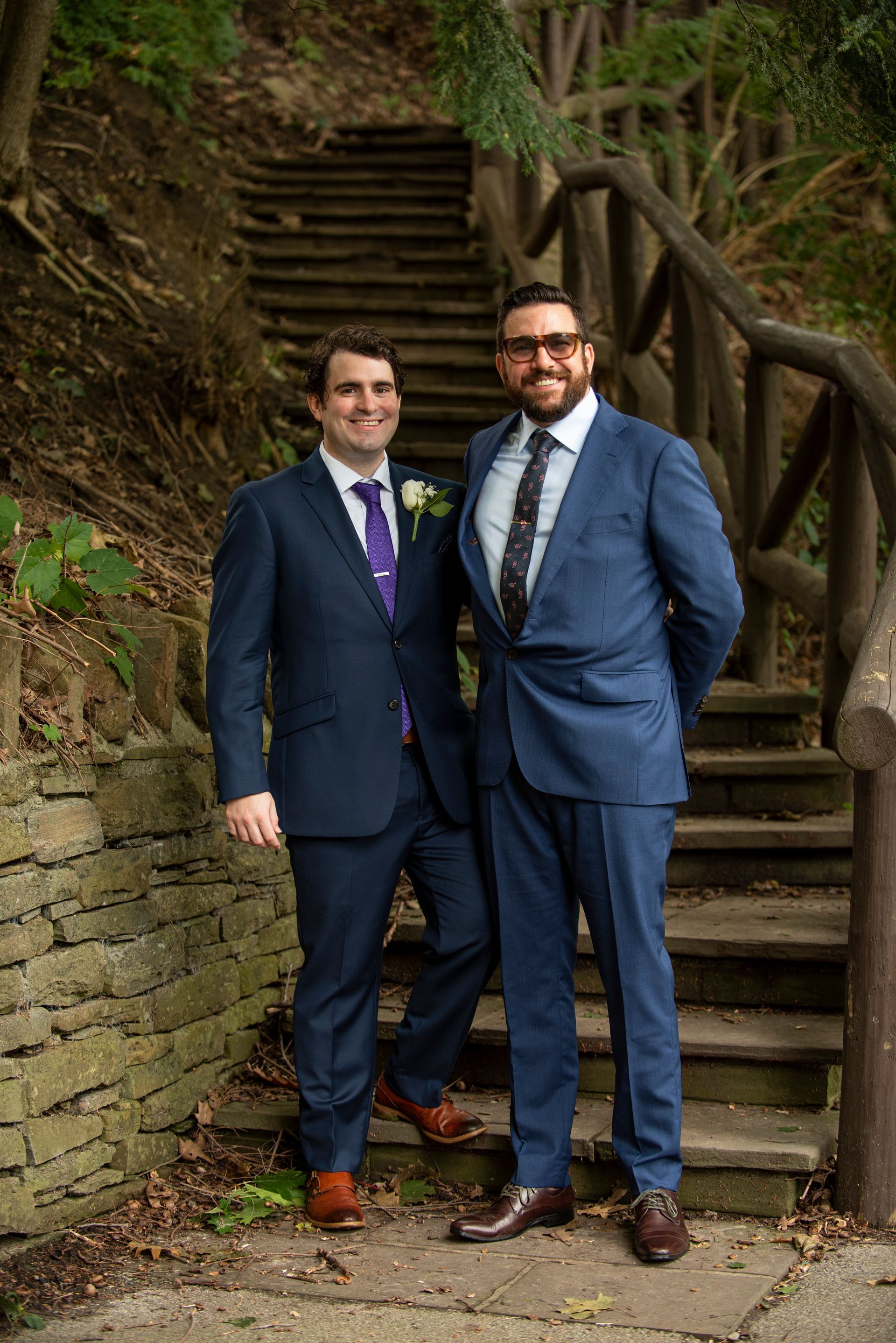 Two men in suits stand on stone steps; one smiles, the other clasps hands behind back. Green foliage surrounds.
