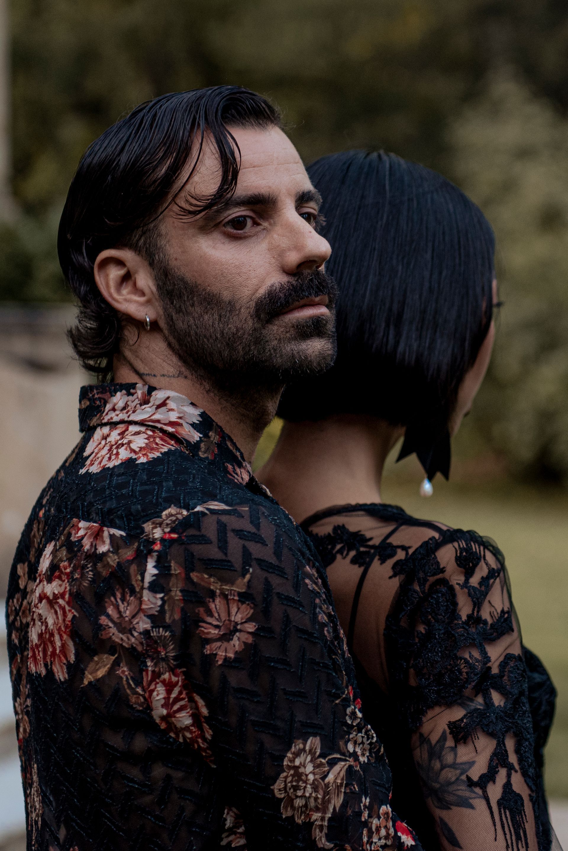 Man in floral shirt, looking away, embraces a person with short black hair. Outdoor setting.