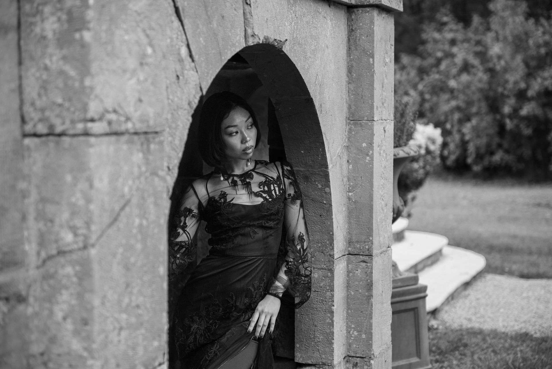 Woman in a black dress poses in a stone archway, gazing to the side. Outdoors with greenery in the background.