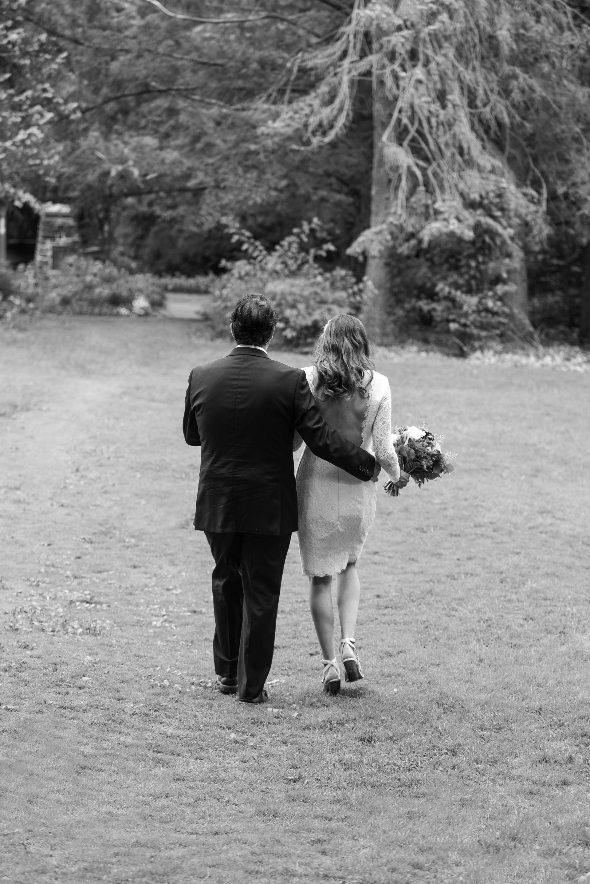 Couple walking away, arm in arm, across a grassy area; holding a bouquet. Trees in background.