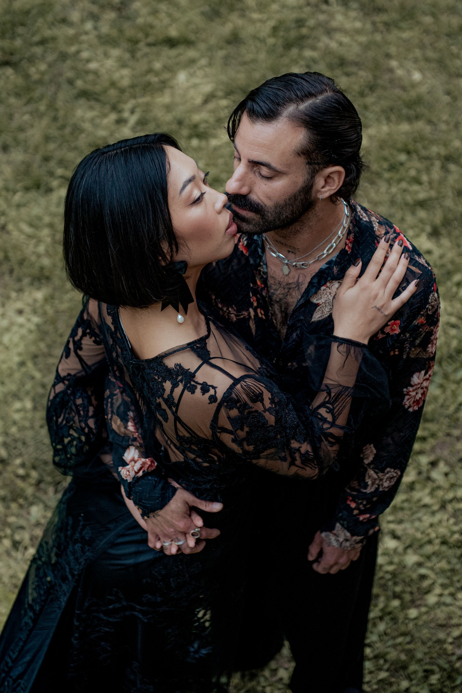 Couple kissing, embraced. Woman in black lace dress, man in floral shirt. Outdoors, green background.
