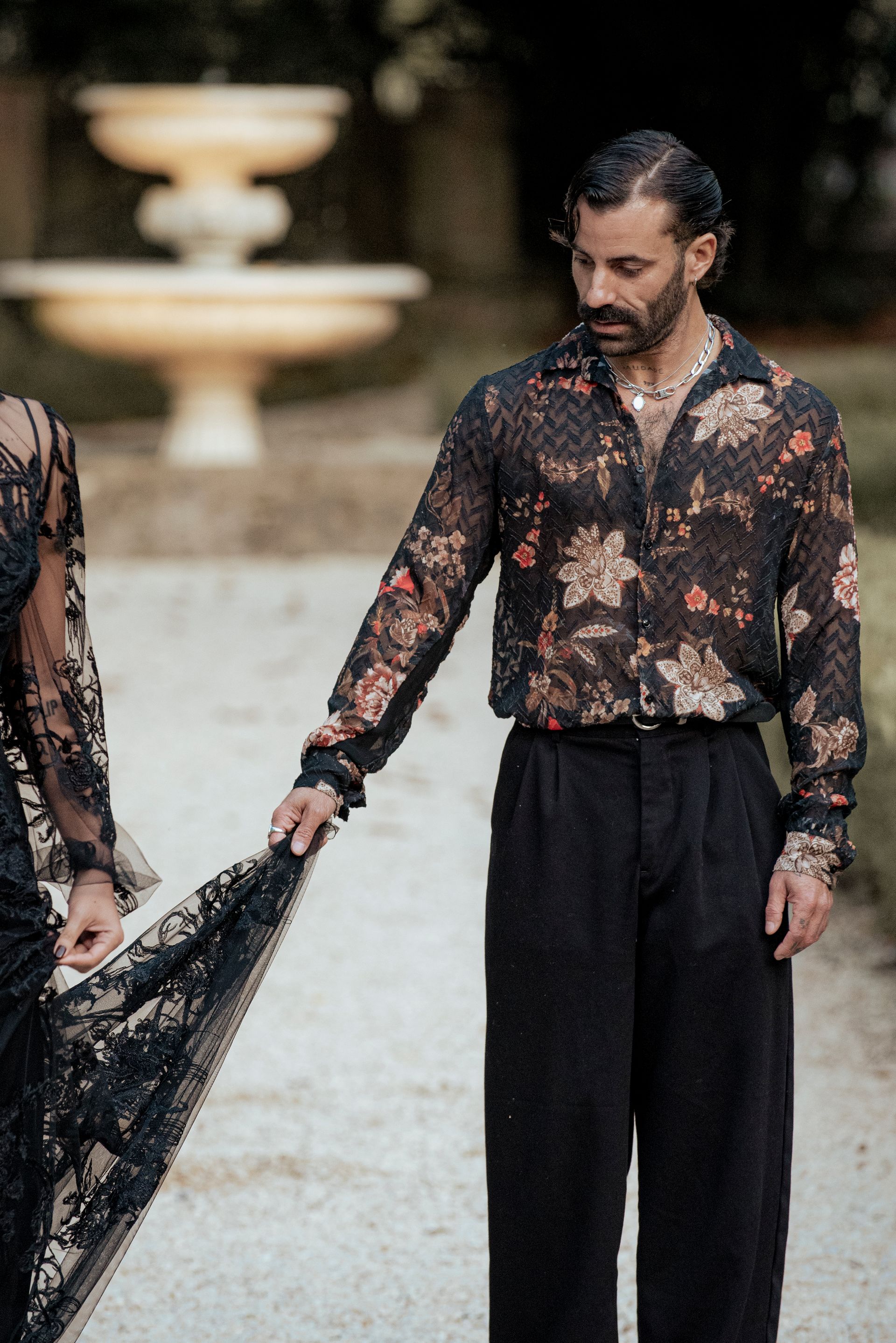 Man in floral shirt holds the lace train of a dress outdoors, near a fountain.