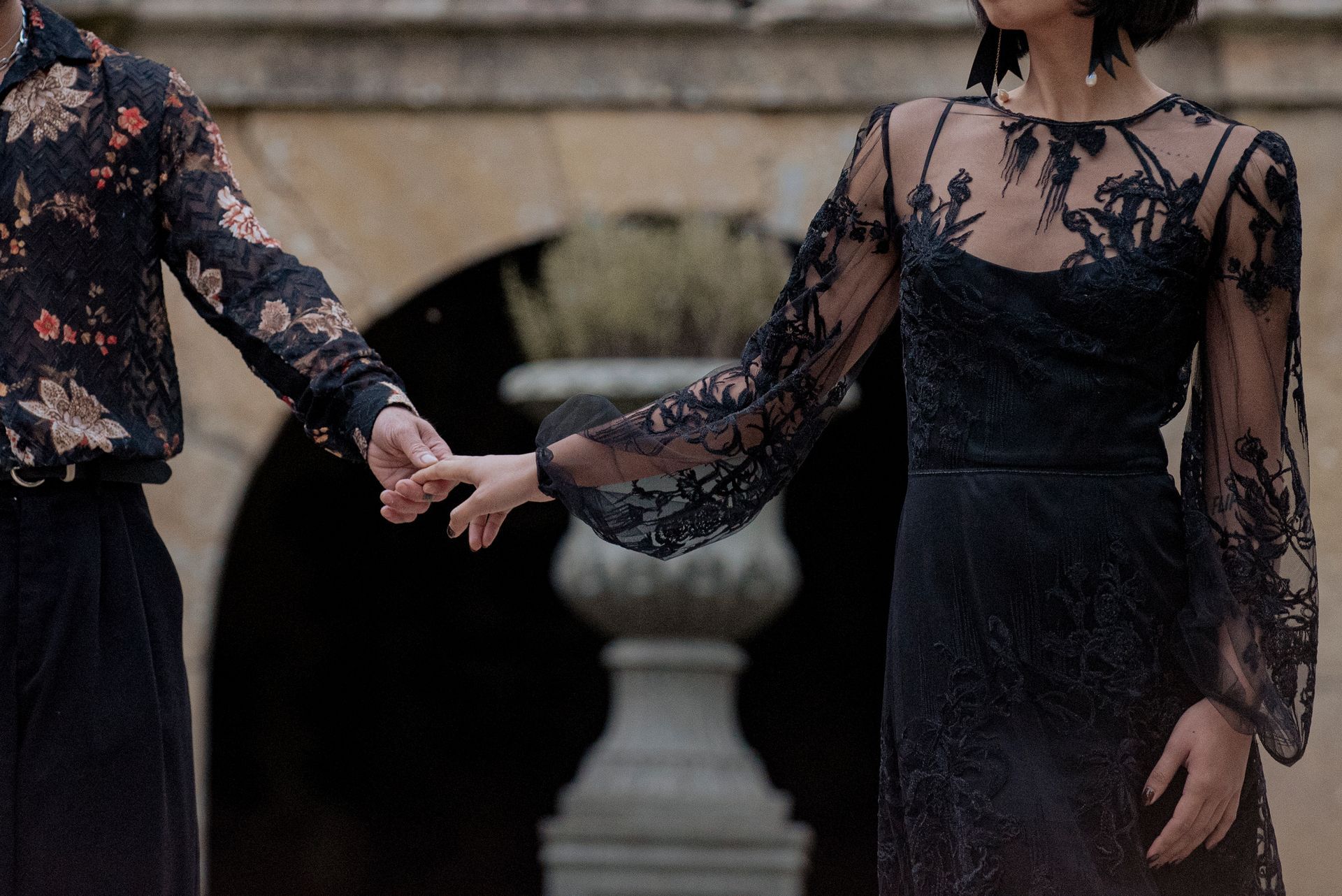 Couple holding hands. Woman in black lace dress, man in floral shirt, outside near stone structure.