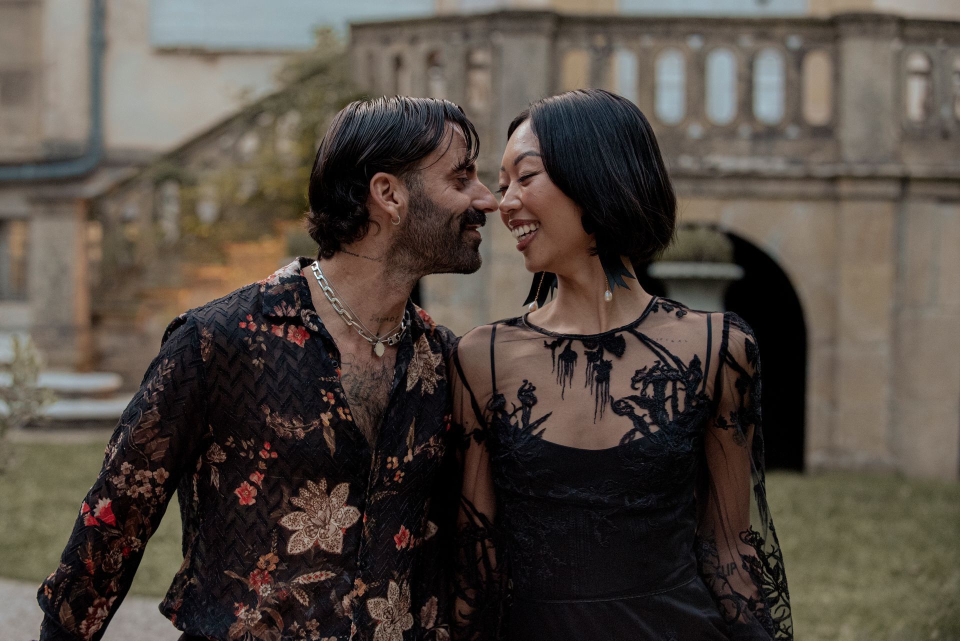 Couple smiles, almost touching noses, outdoors near a castle. They wear black formal wear.