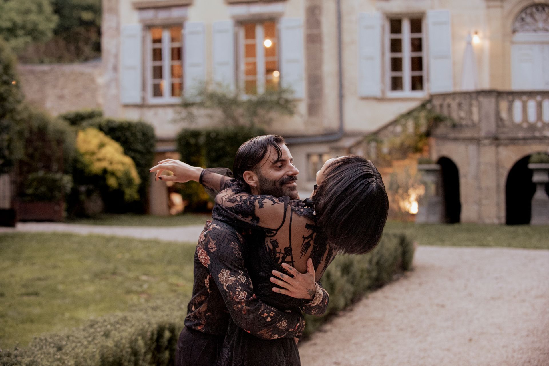 Couple embracing outdoors near a large building, smiling. Evening, soft light.