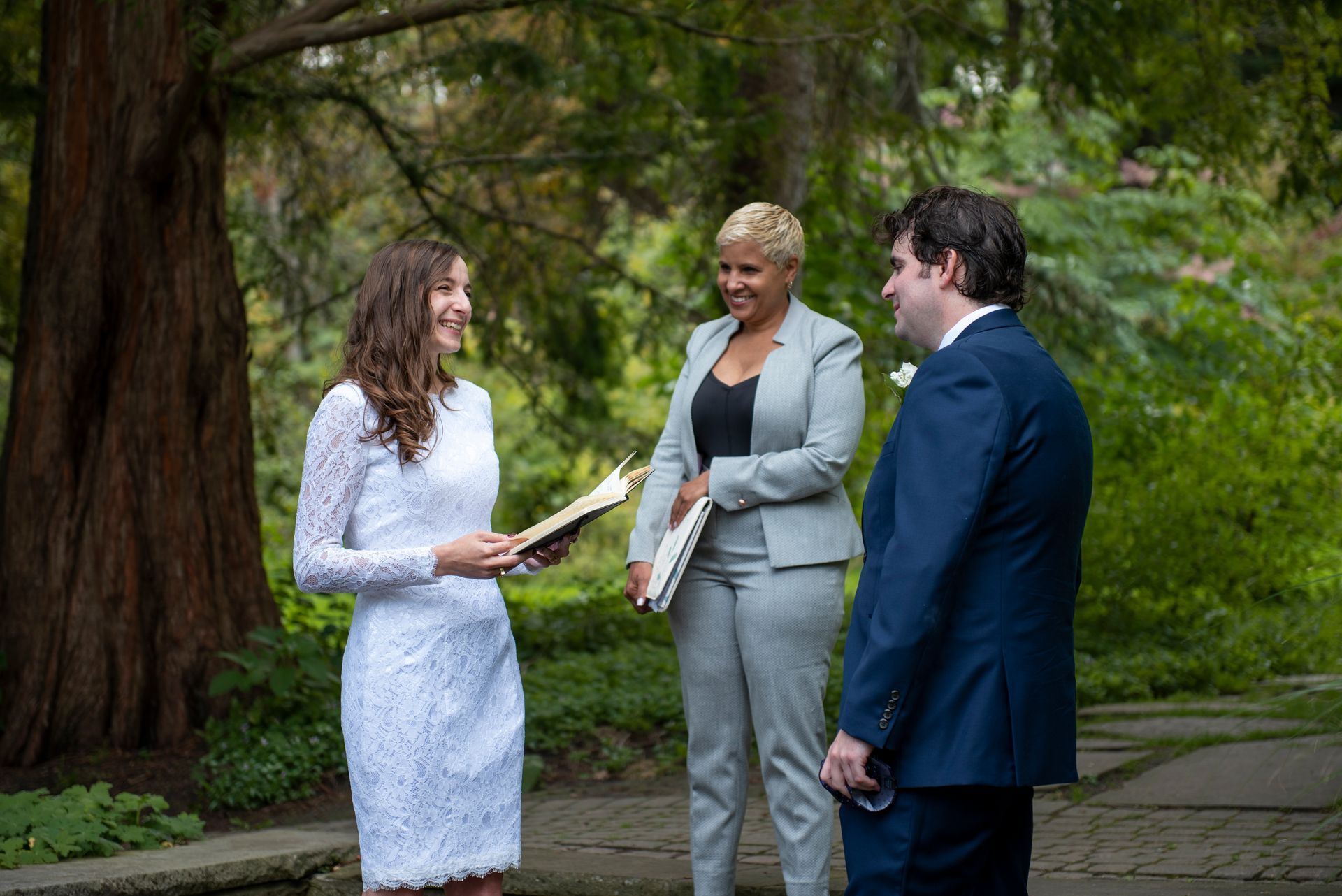 Wedding ceremony outdoors: couple with officiant under trees. Woman in white dress smiles; the man in a blue suit smiles. Officiant wears a gray suit.