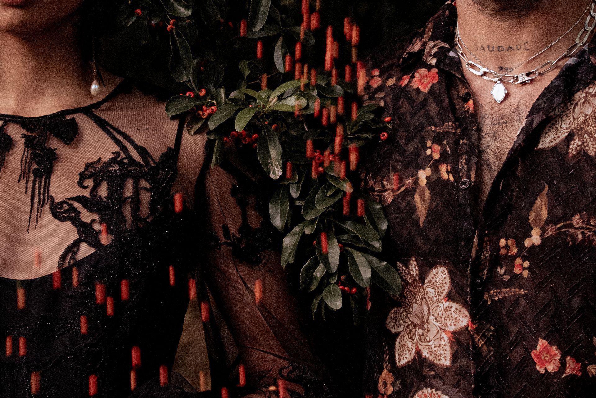 Close-up of a couple, partially obscured by foliage. Man in floral shirt, woman in black lace dress. Rain effect.