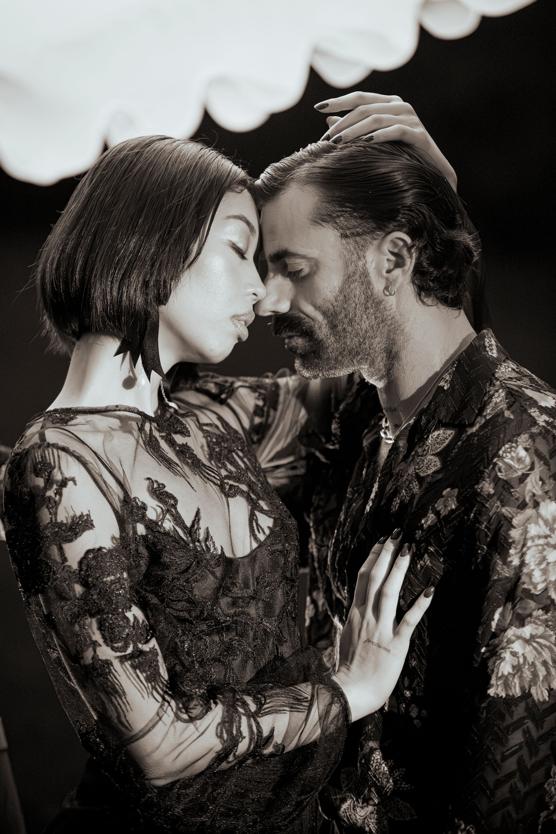 Couple in embrace. She wears sheer, black dress; he touches her hair.