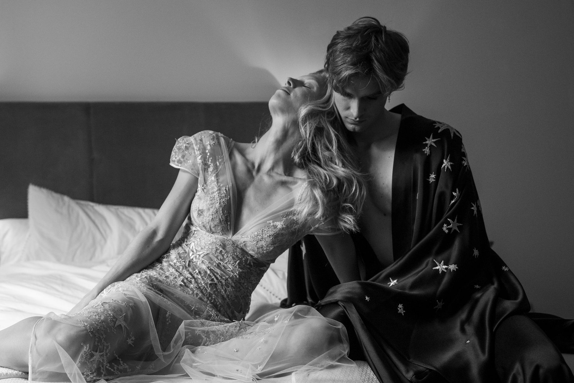 Woman in embellished gown leans back on bed with man in robe, intimate setting.