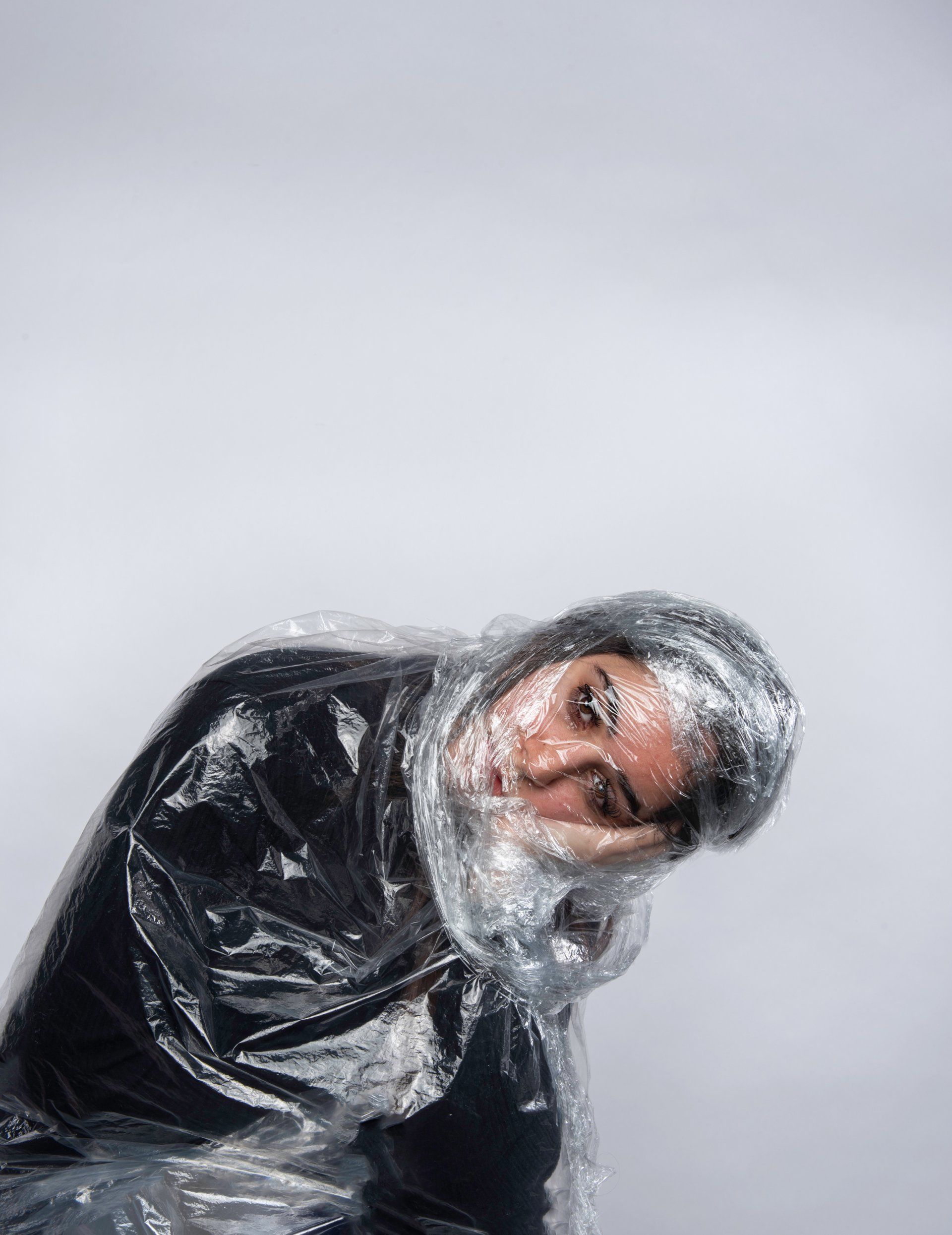 Person wrapped in clear plastic, head tilted, against a white background.