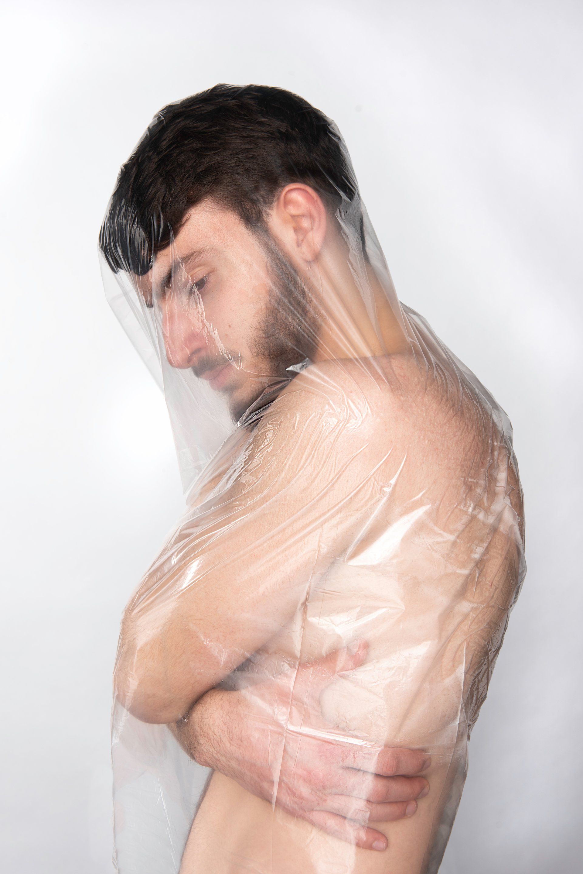 Man wrapped in clear plastic, arms crossed, looking down. Against a white background.