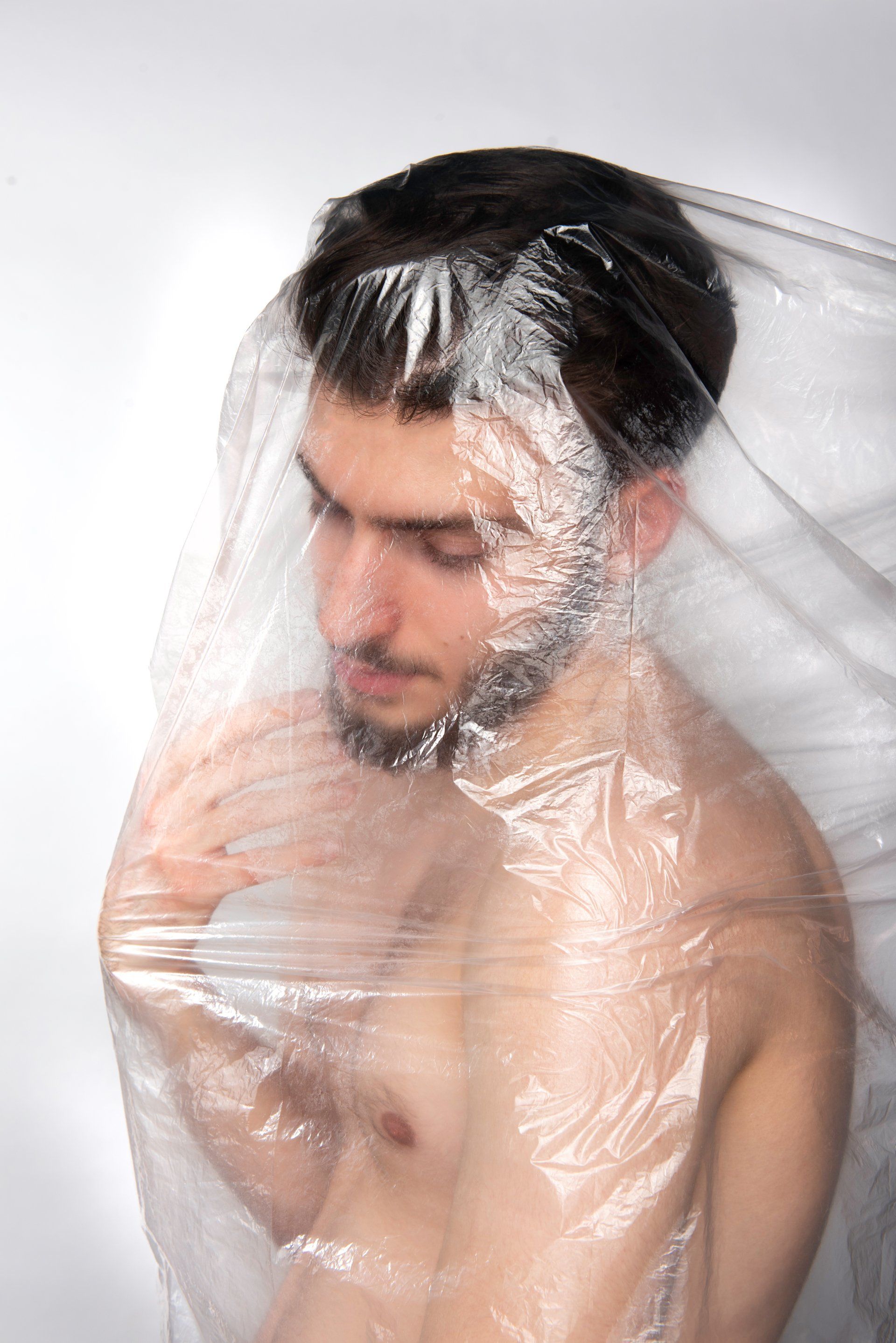 Man with dark hair and beard, partially covered in translucent plastic sheeting, looking down.