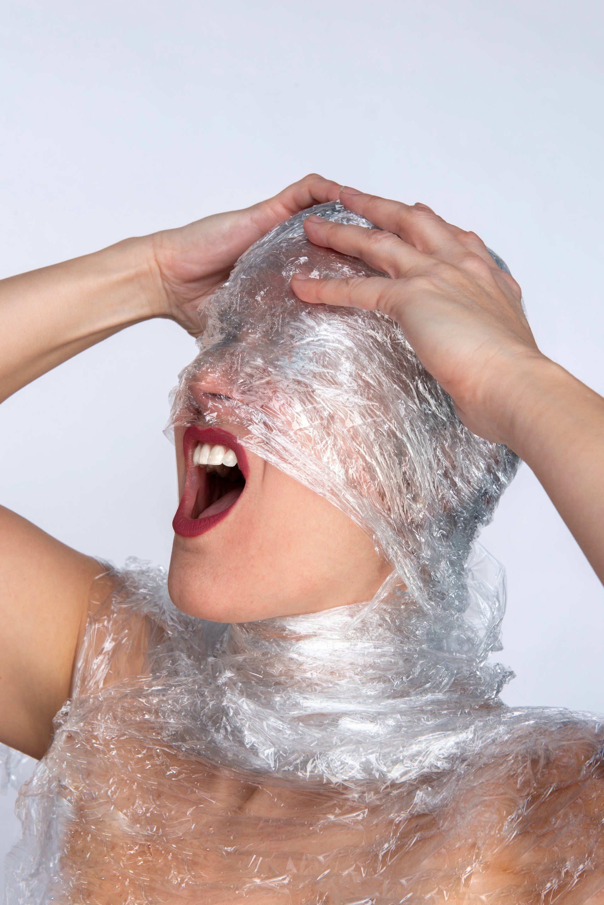 Woman's head and neck wrapped in plastic. She's screaming, hands on head, against a white background.