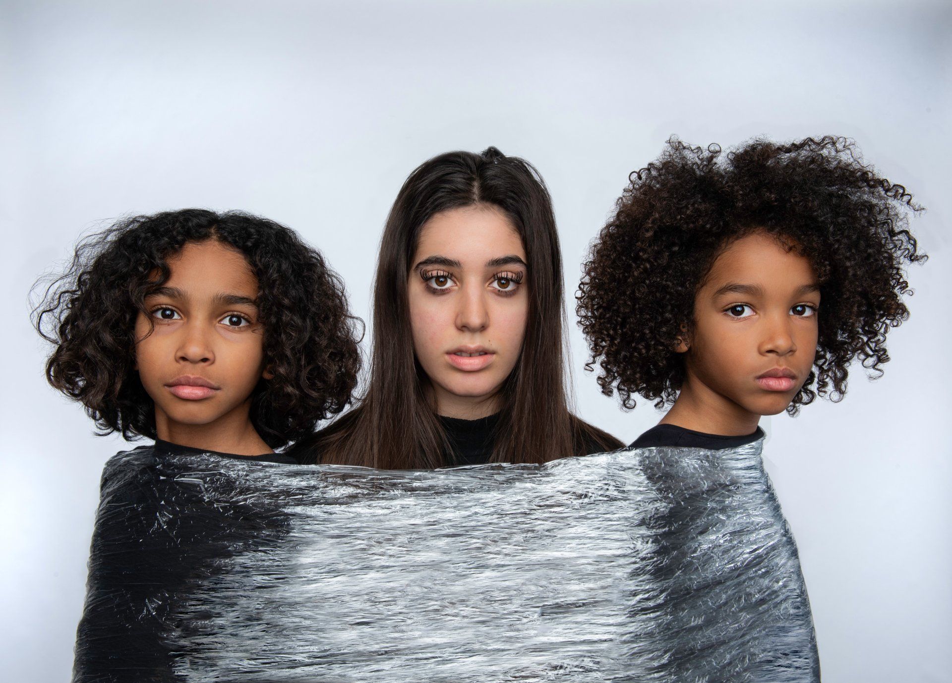 Three people: one in the center, and two on the sides, partially wrapped in a silver-painted textile, all looking at the camera.