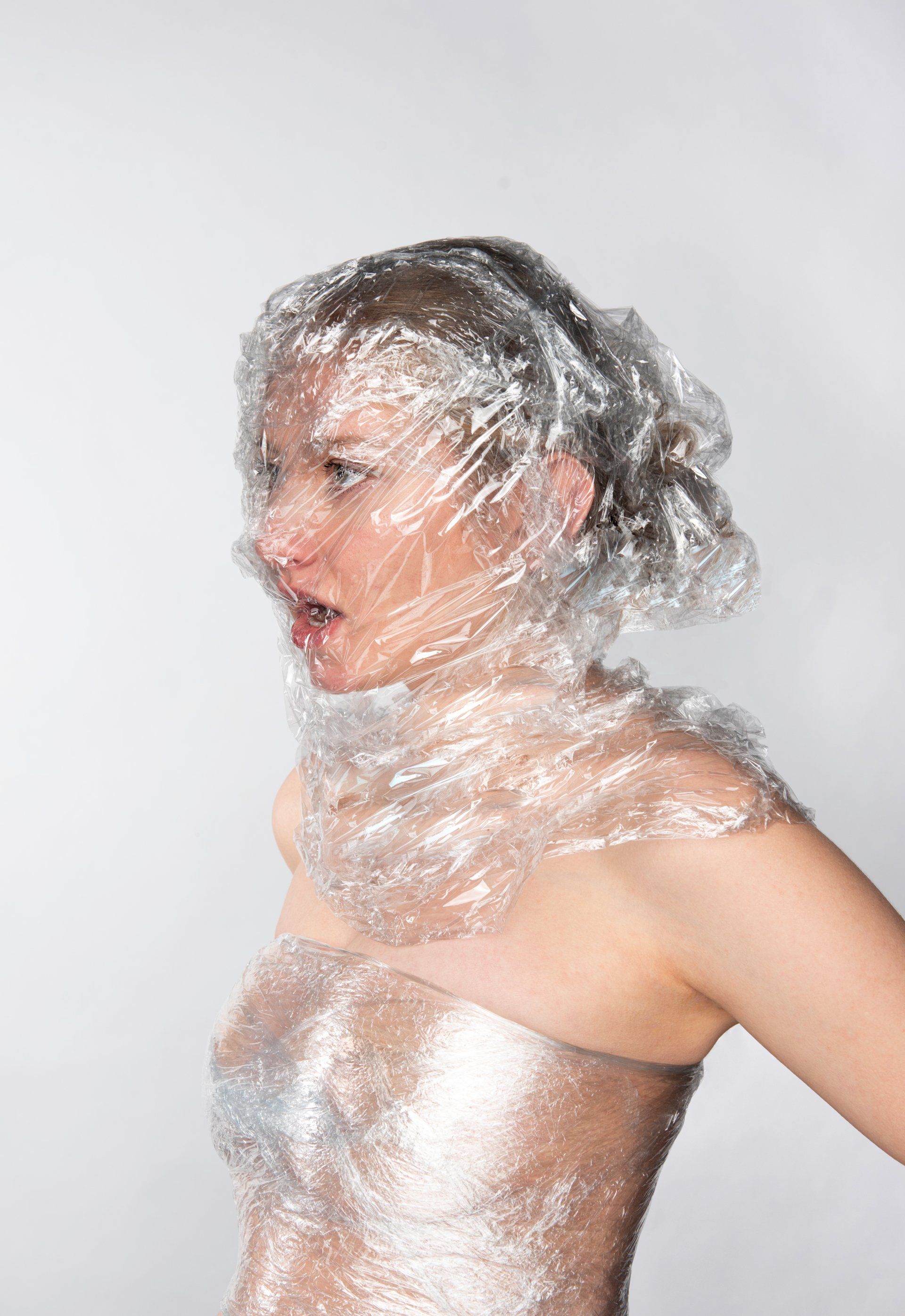 Woman wrapped in clear plastic, torso exposed, mouth open, looking left against white background.