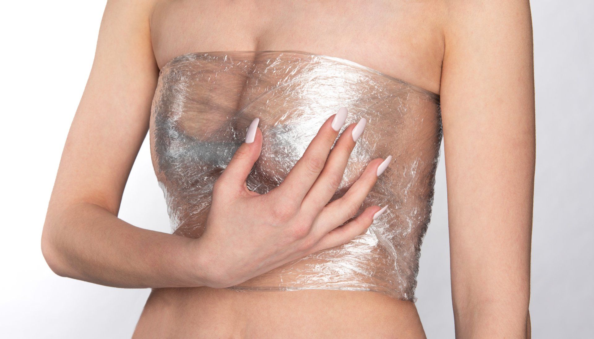 Woman wrapped in clear plastic wrap, touching chest area, against a white background.