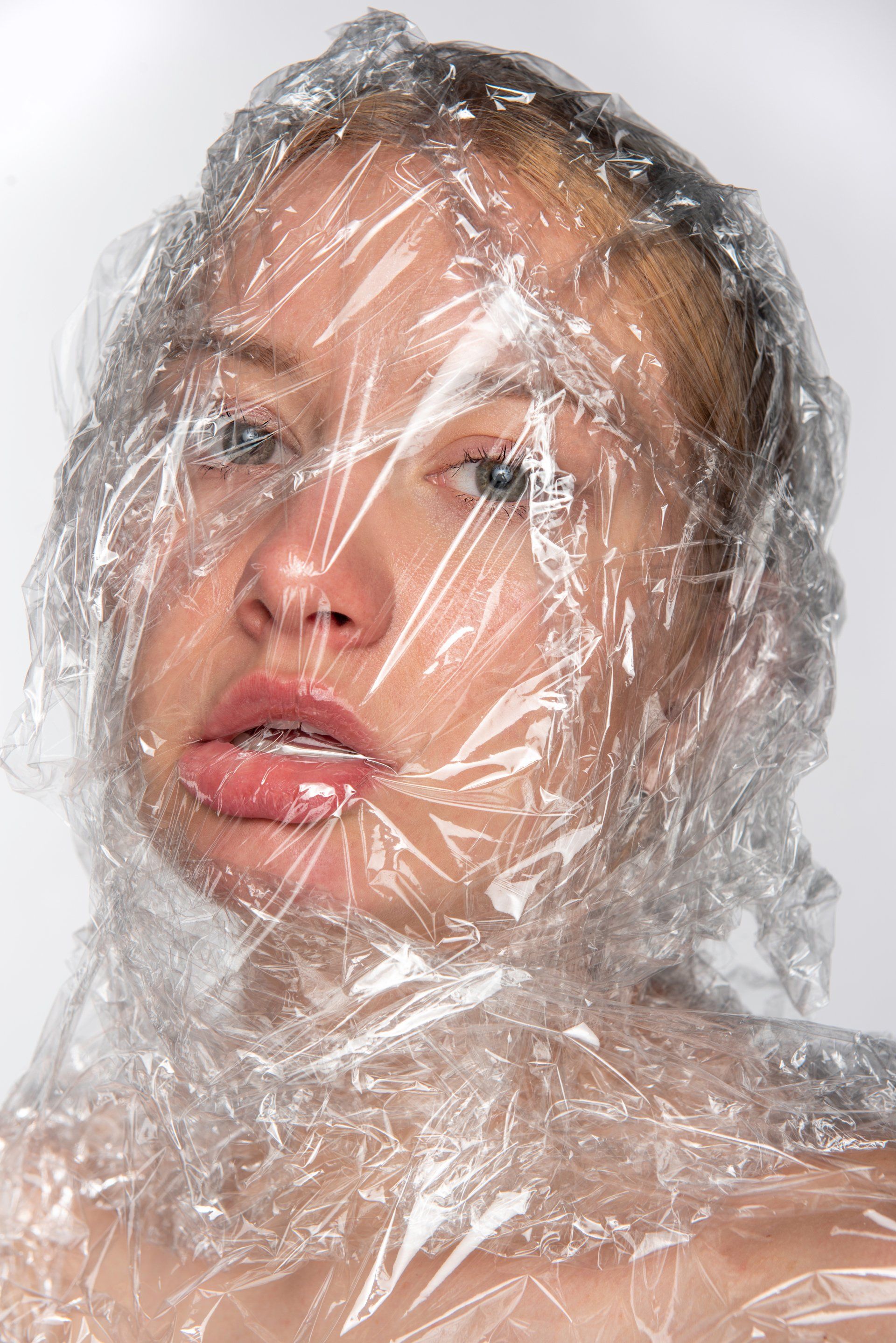 Woman's face wrapped in clear plastic, looking directly at the viewer with a neutral expression.