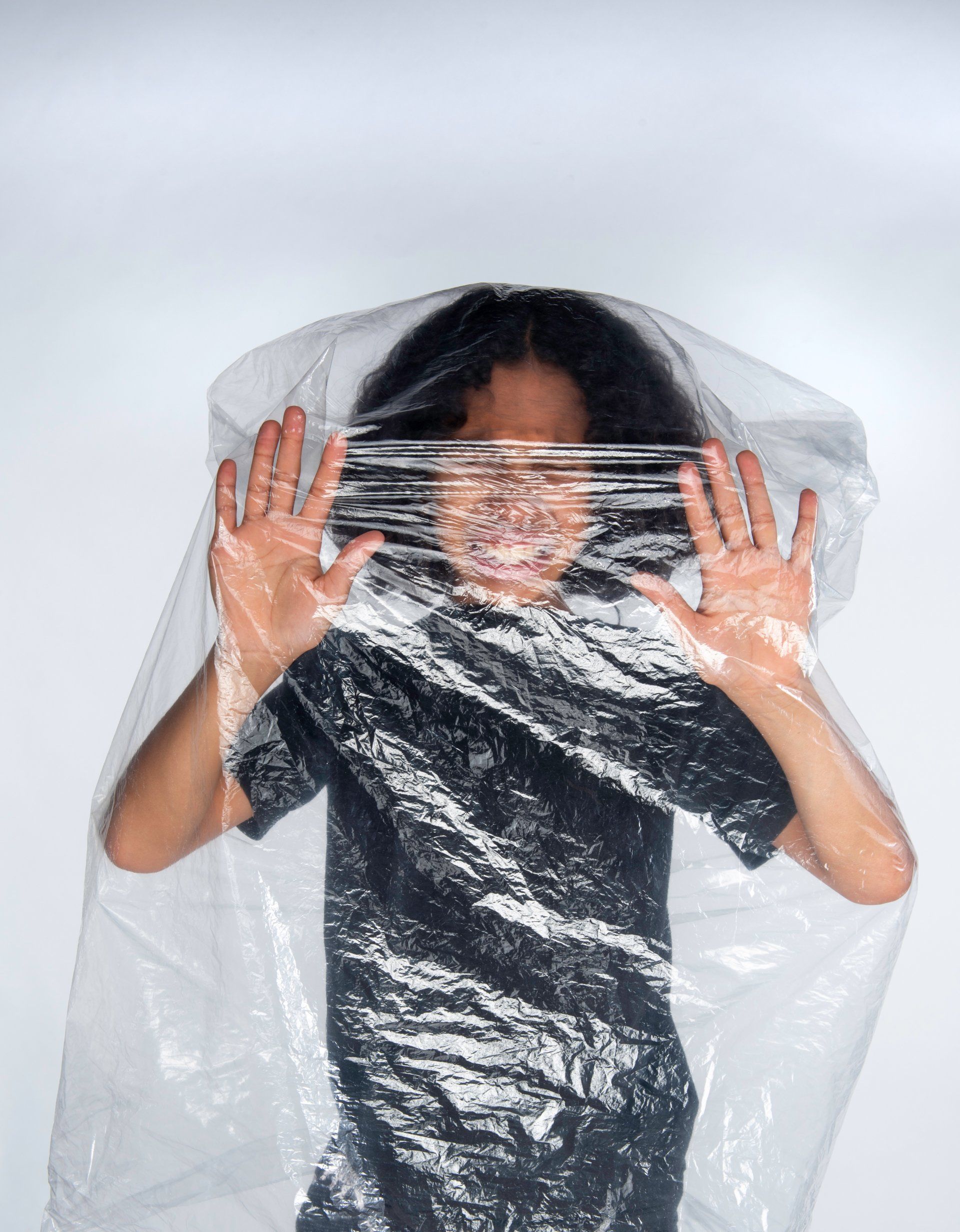 Person with hands pressed against clear plastic. Black shirt, wavy hair, white background.