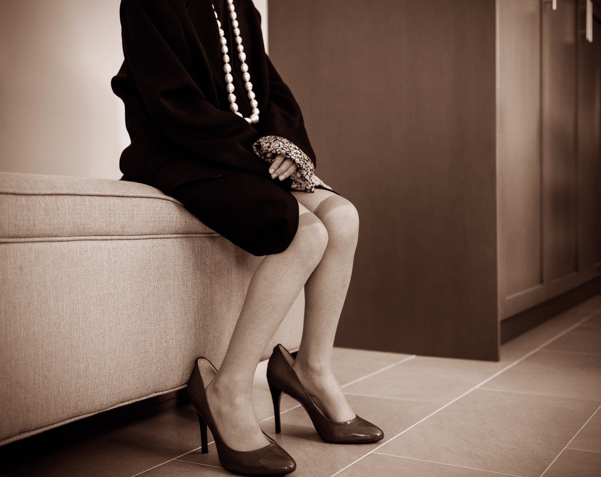 Woman seated on a bench, wearing a black outfit, pearl necklace, and high heels.