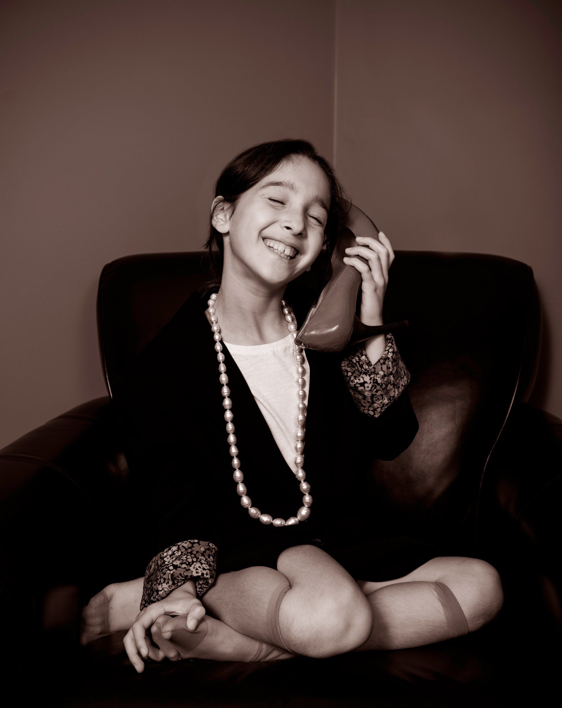 Girl in oversized clothes, holding a high-heeled shoe to her ear, smiling in a leather chair.