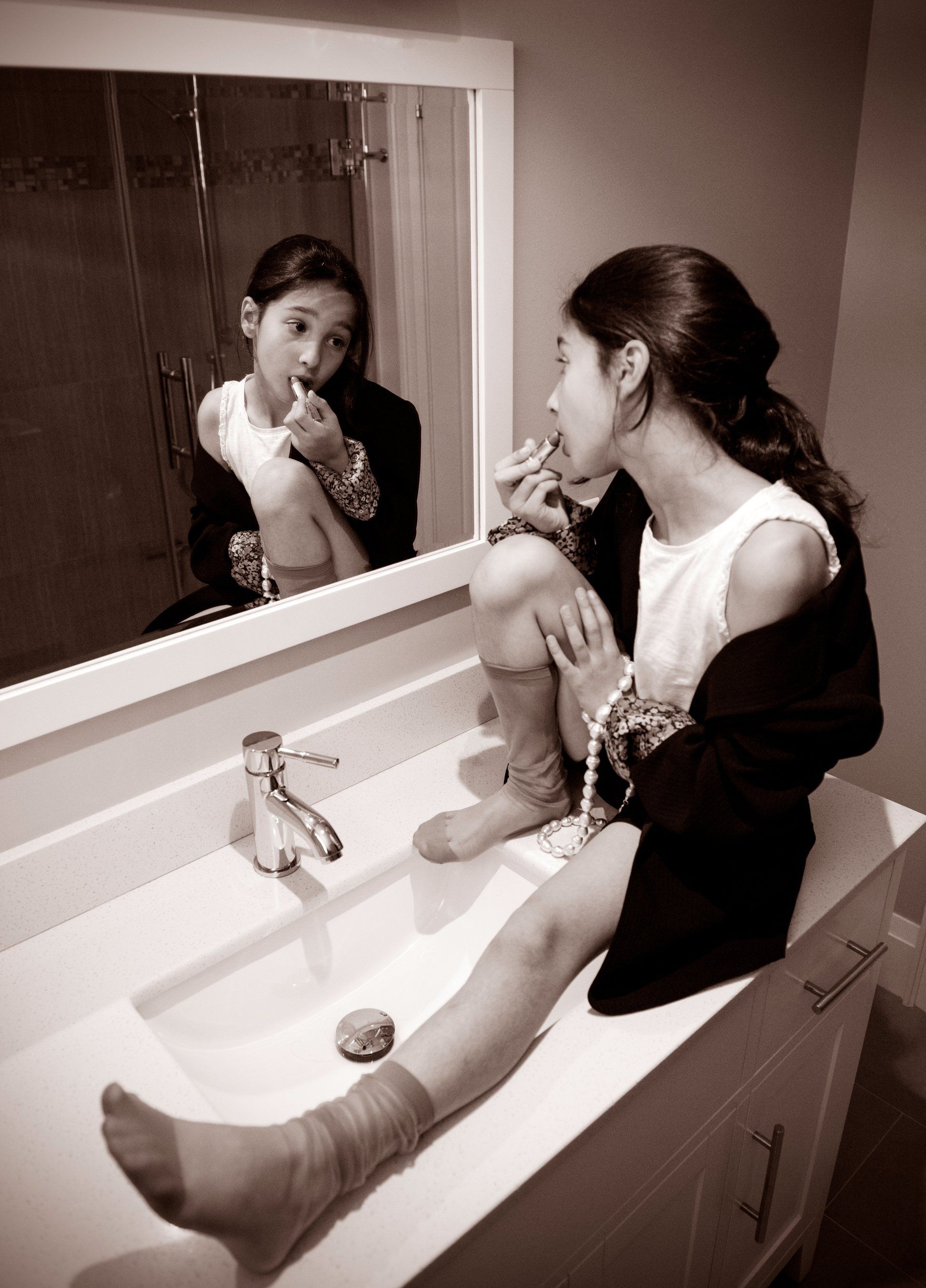 Girl seated on a bathroom sink applies lipstick, looking in a mirror. Wearing a blazer, t-shirt, socks.