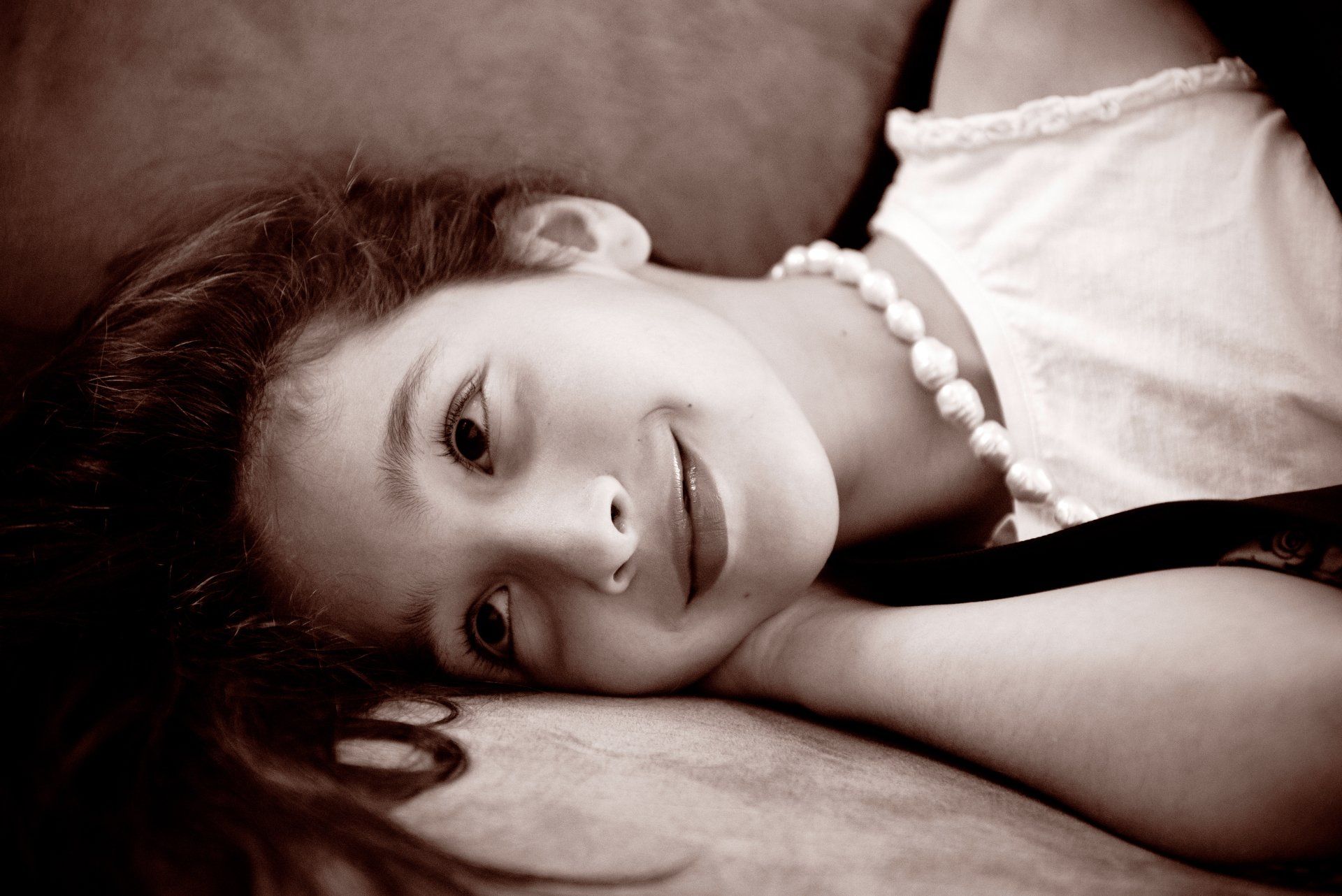 Girl smiling, laying on a couch, wearing a white top and necklace. Sepia tone.