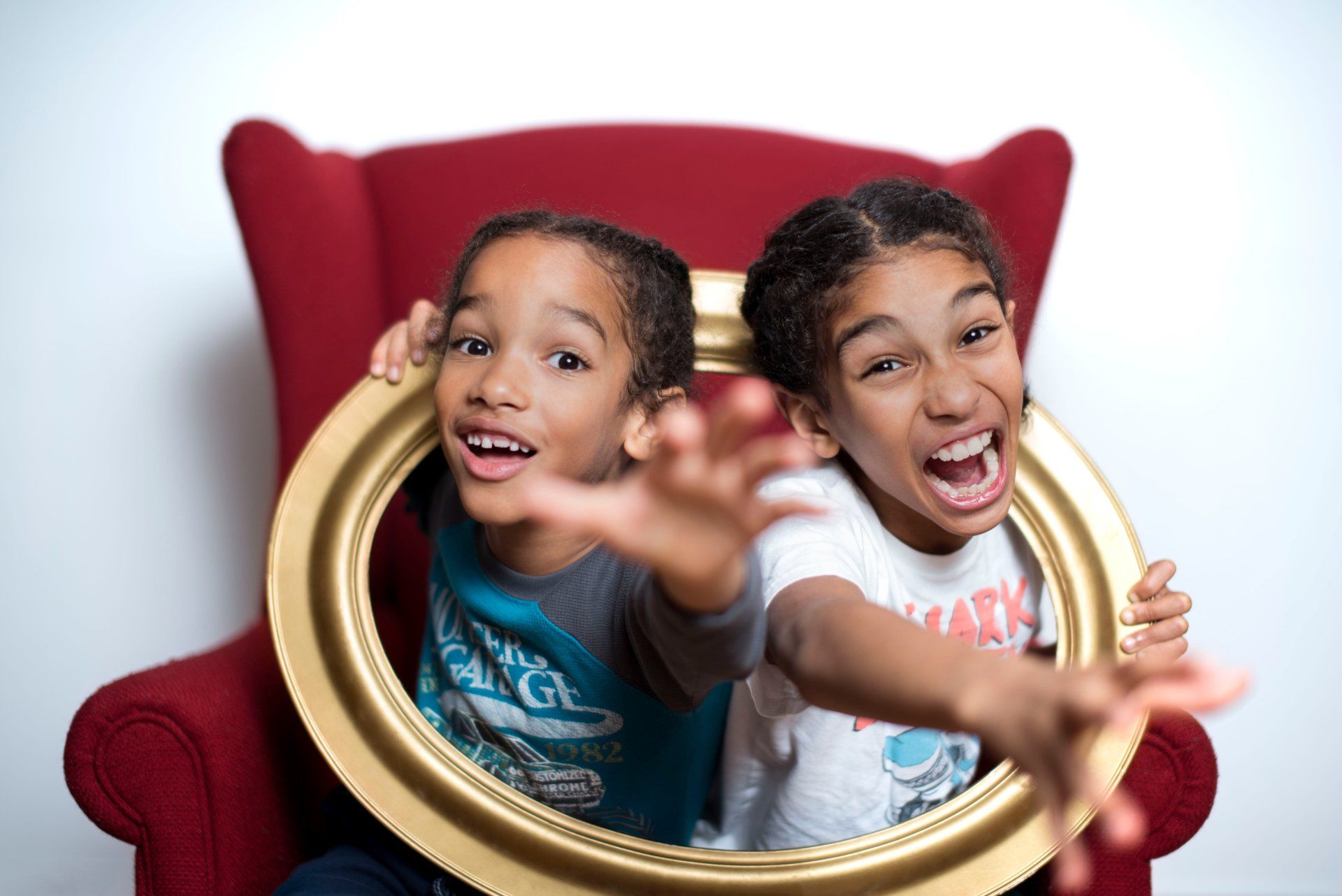 Two children reaching out through a gold picture frame, sitting in a red armchair, against a white background.