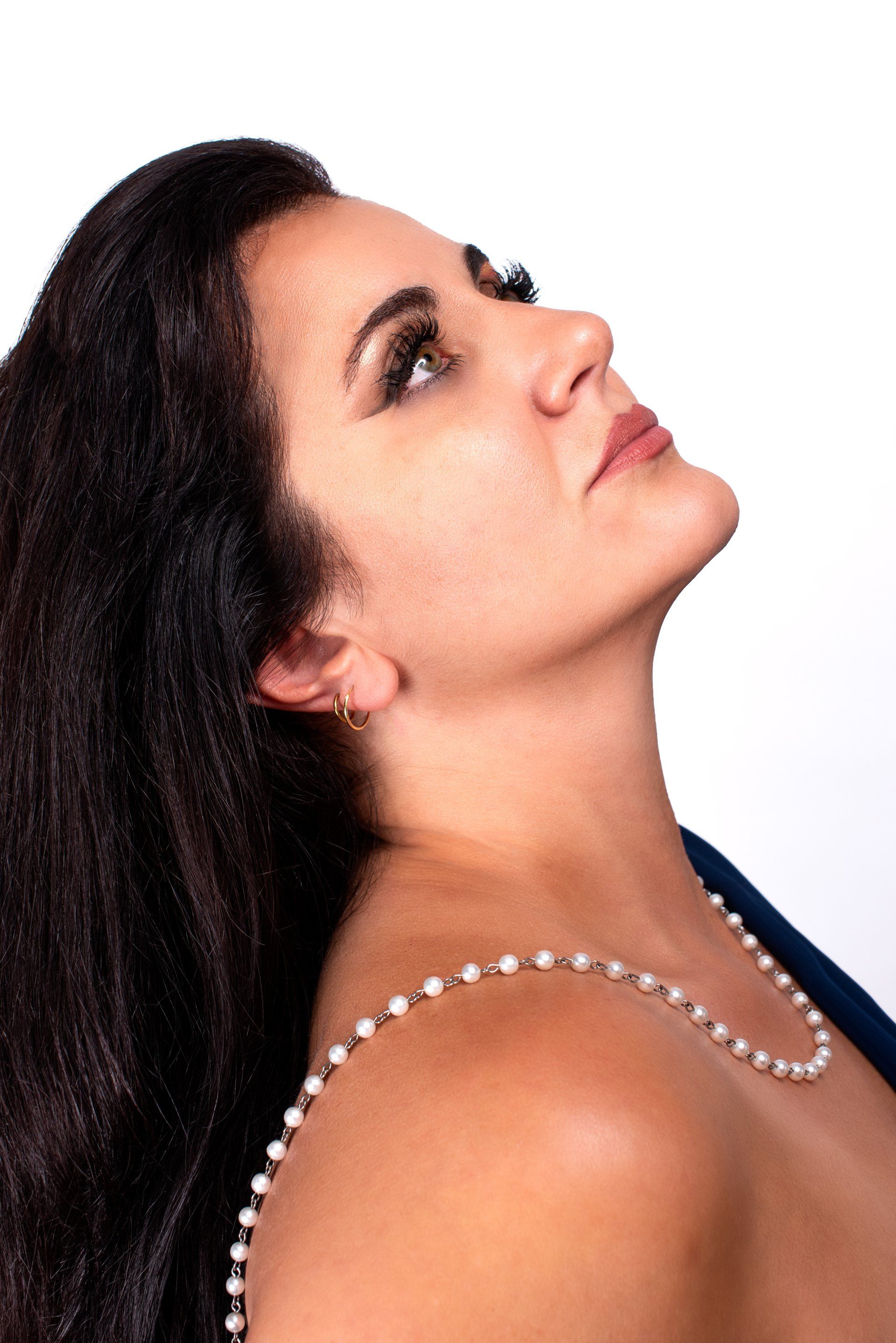 Woman looking upward, wearing a pearl necklace and a blue jacket. Dark hair.