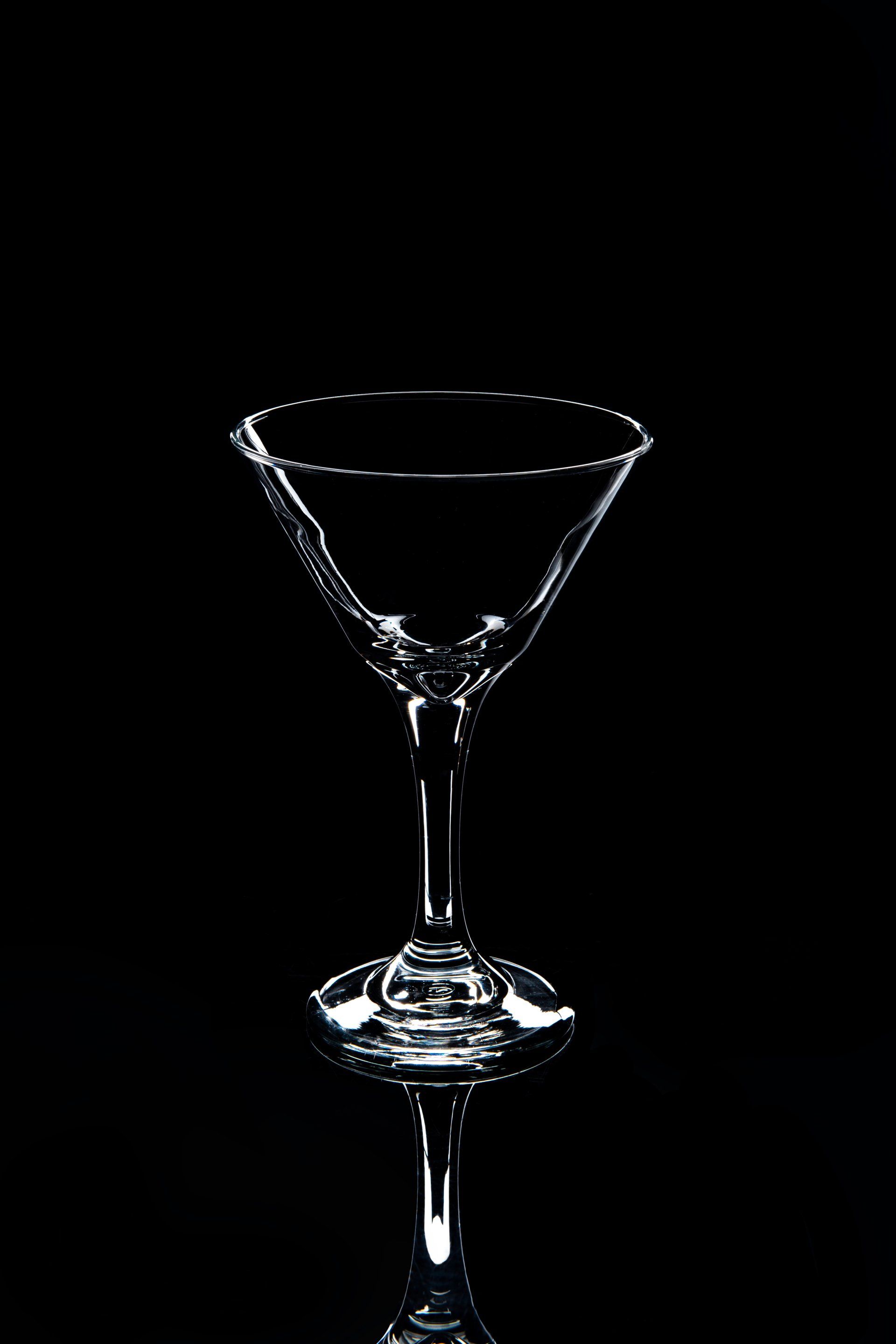Empty martini glass, clear with a long stem, on a reflective black surface.