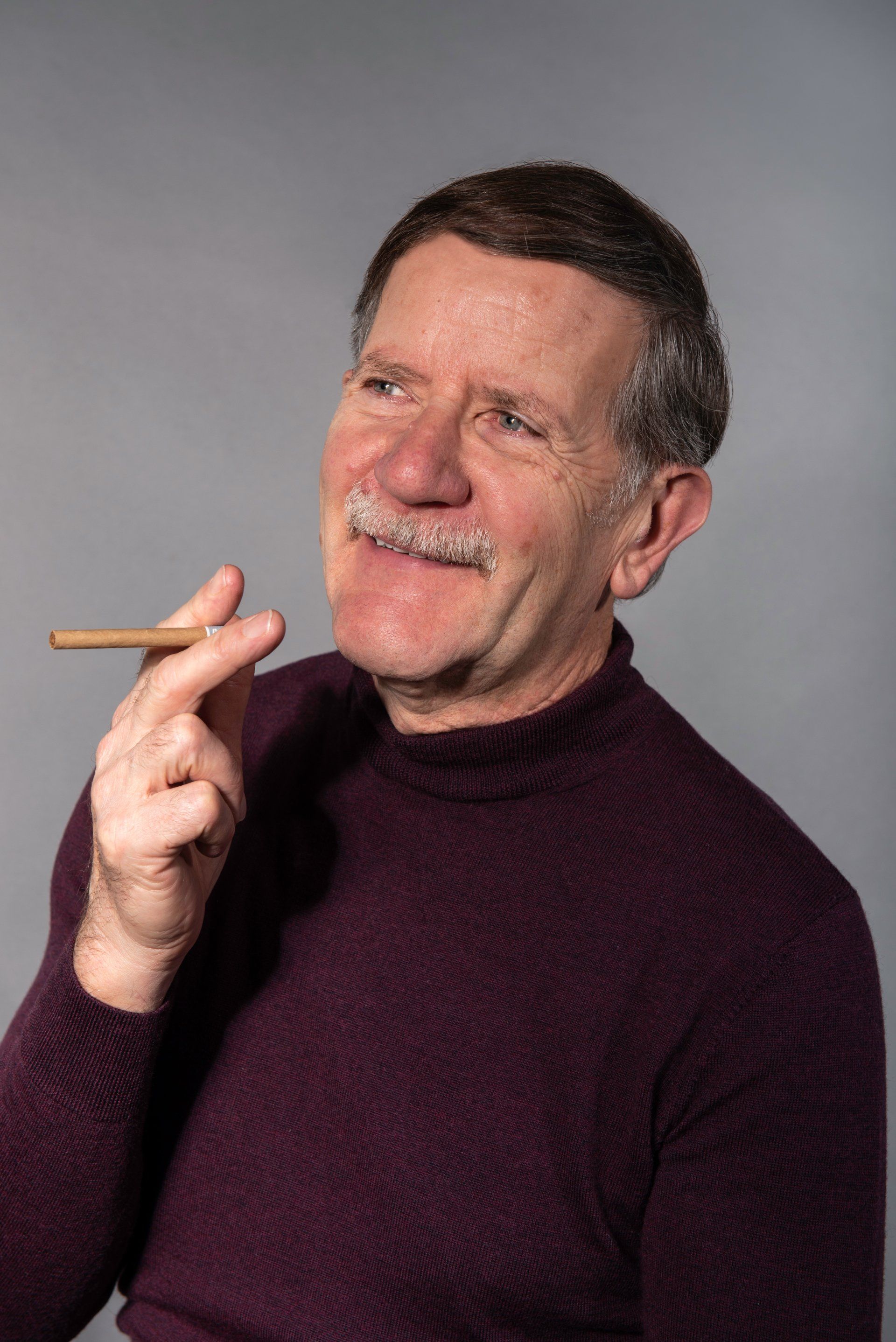 Man with mustache holds lit cigar, looking up and to the right; wearing a burgundy turtleneck.
