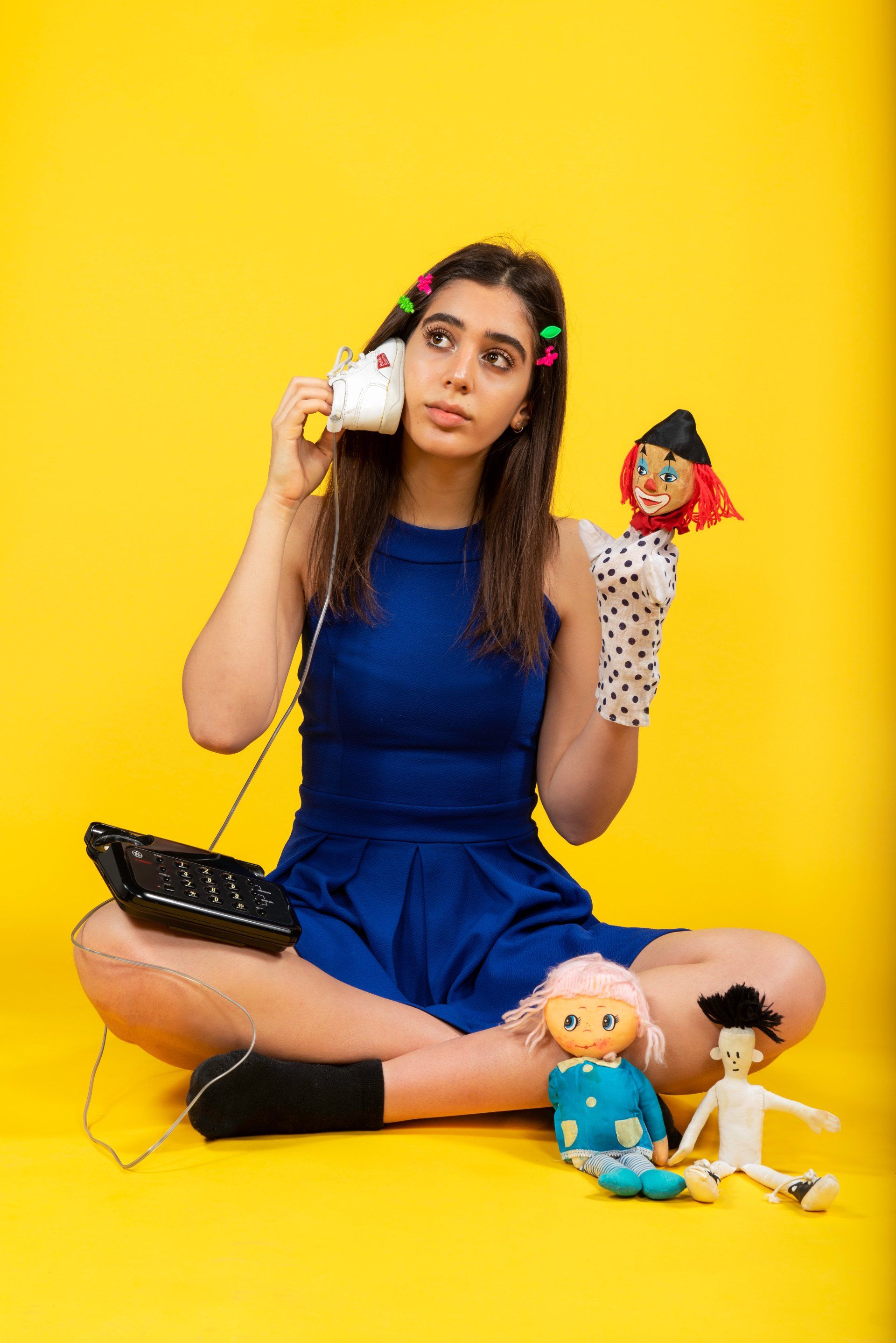 Woman on yellow background holding a phone and puppet, with dolls nearby. She wears a blue dress and stares upwards.