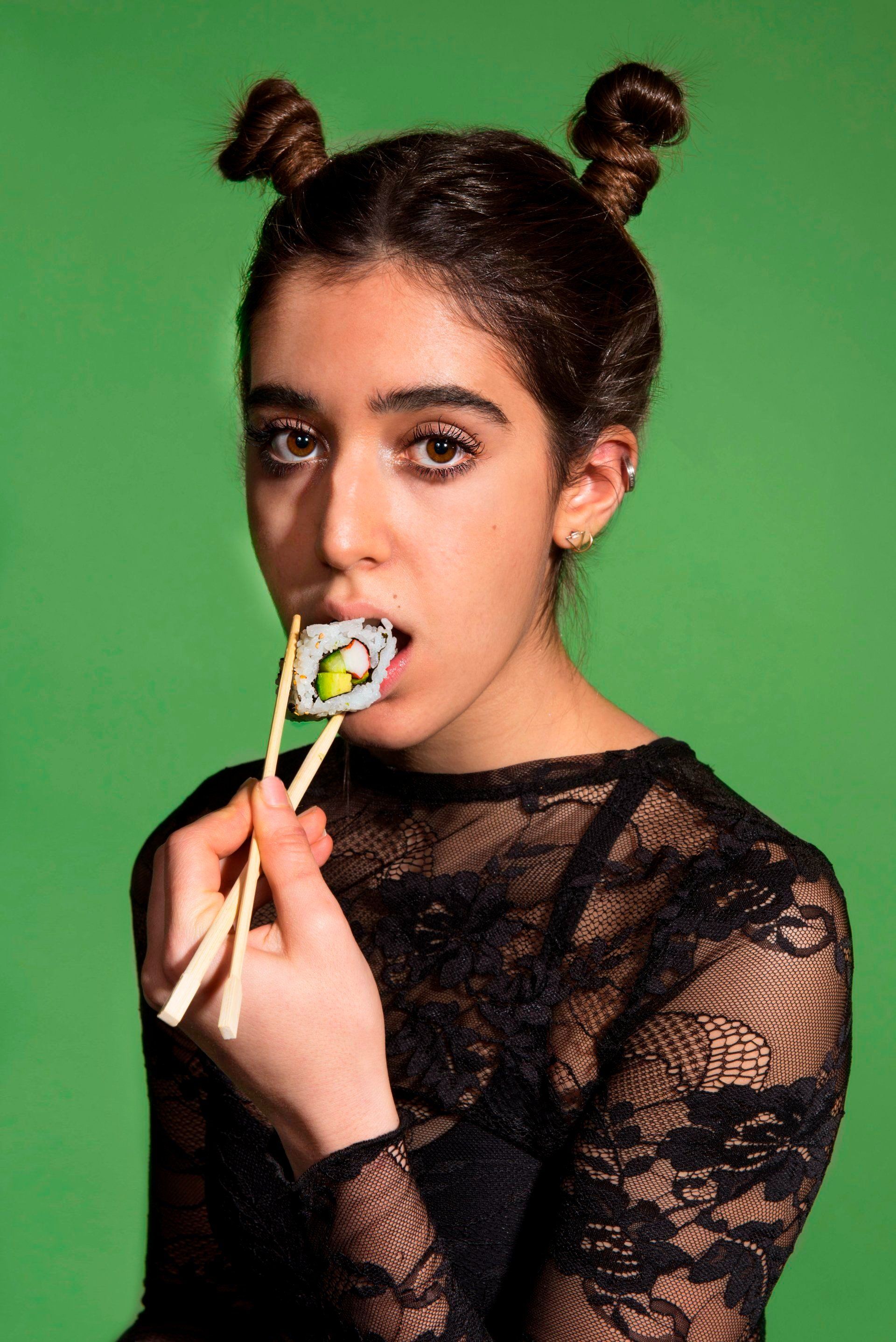 Woman with space buns eating sushi from chopsticks, green background.