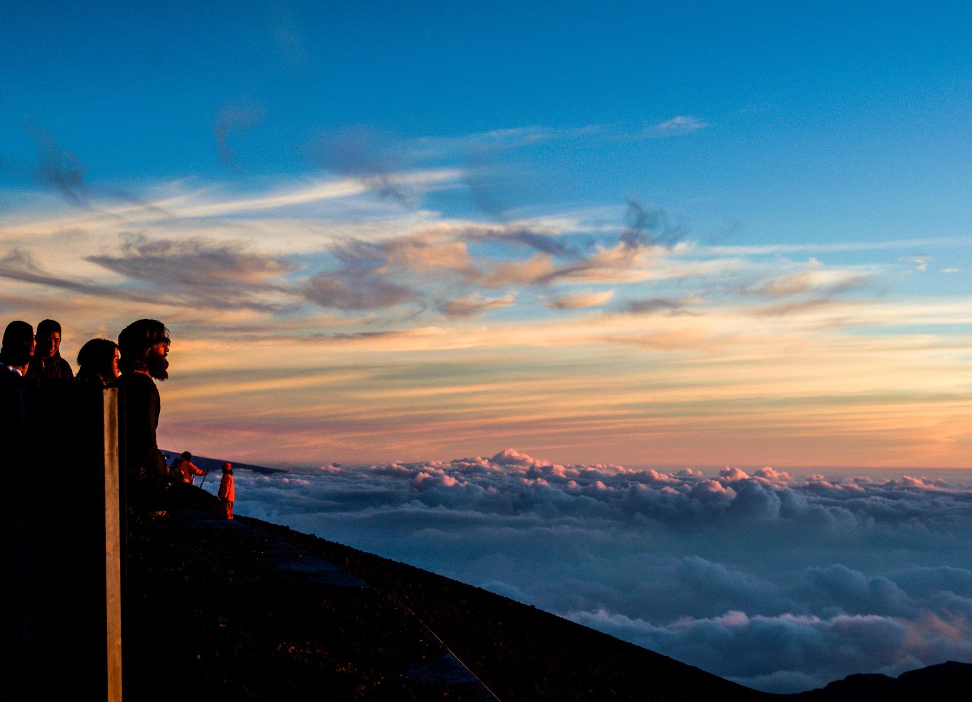 People silhouetted, watching sunrise over clouds. Blue and orange sky.