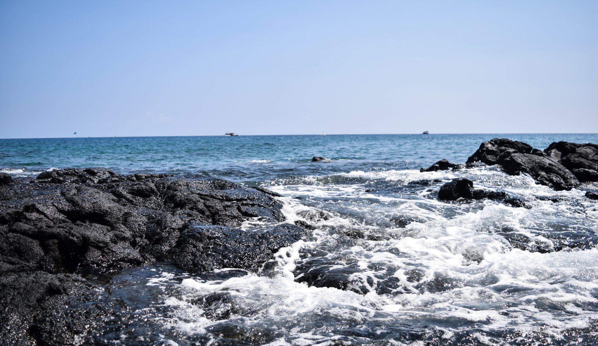 Waves crashing against dark rocks at a sunny beach. Blue sky and ocean in the background.