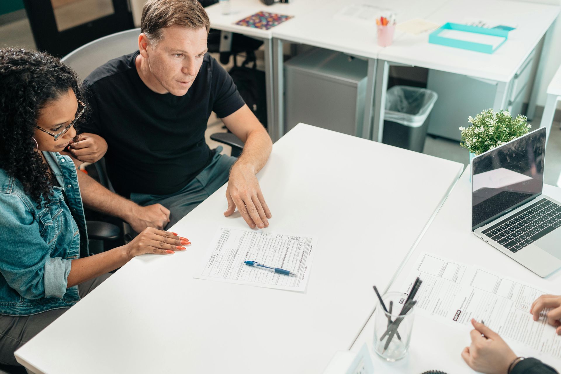 Two people reviewing papers at a white table with a laptop in a bright office setting.