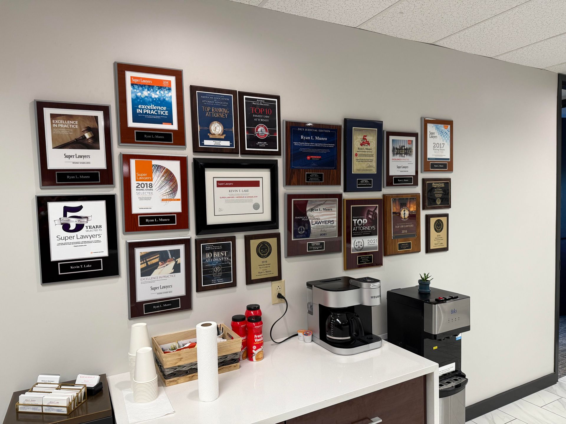 Wall with framed awards, coffee station with supplies, and a water dispenser in an office.